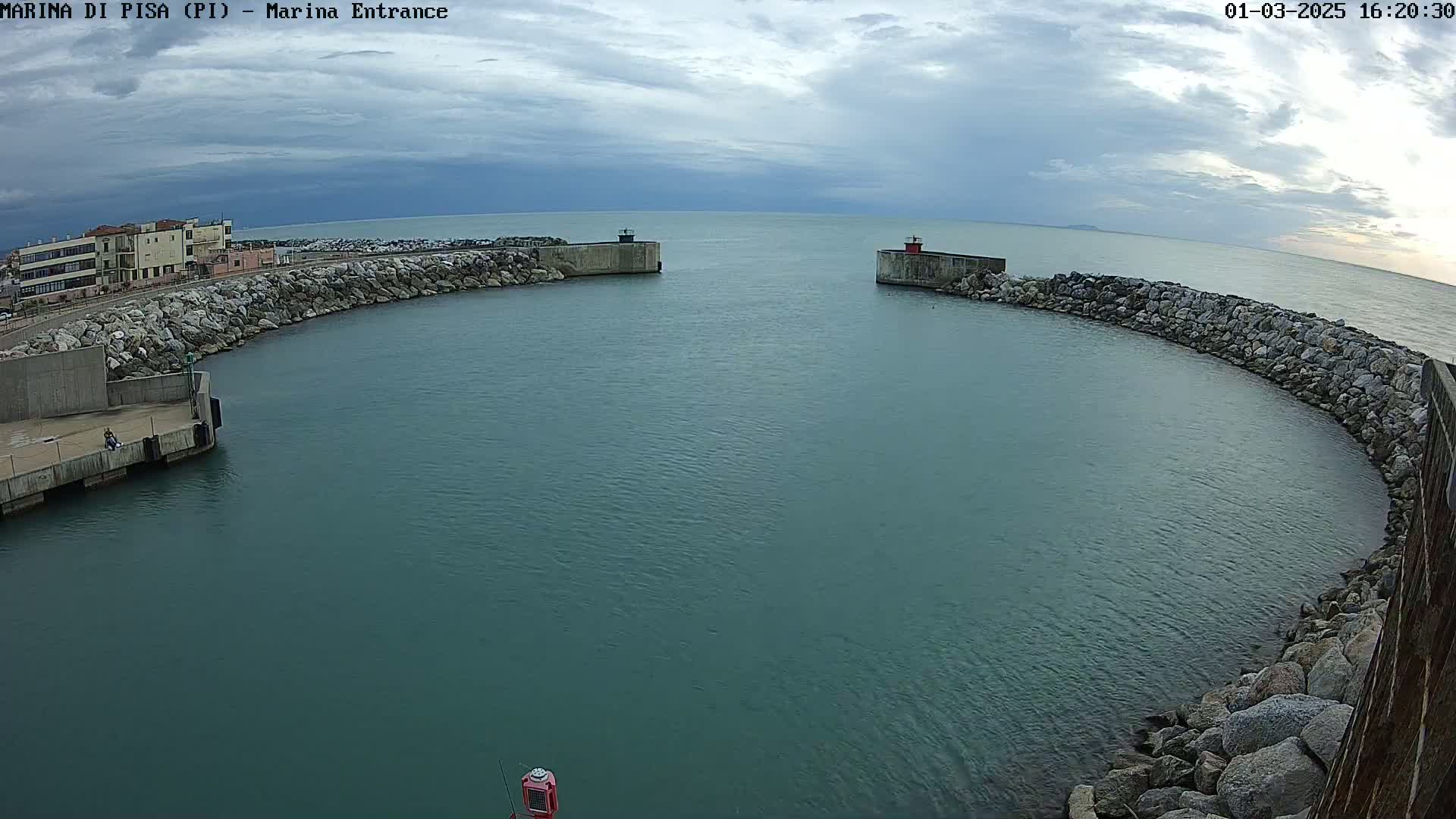 A calm, teal-colored harbor is partially enclosed by gray stone walls under a mostly cloudy sky.