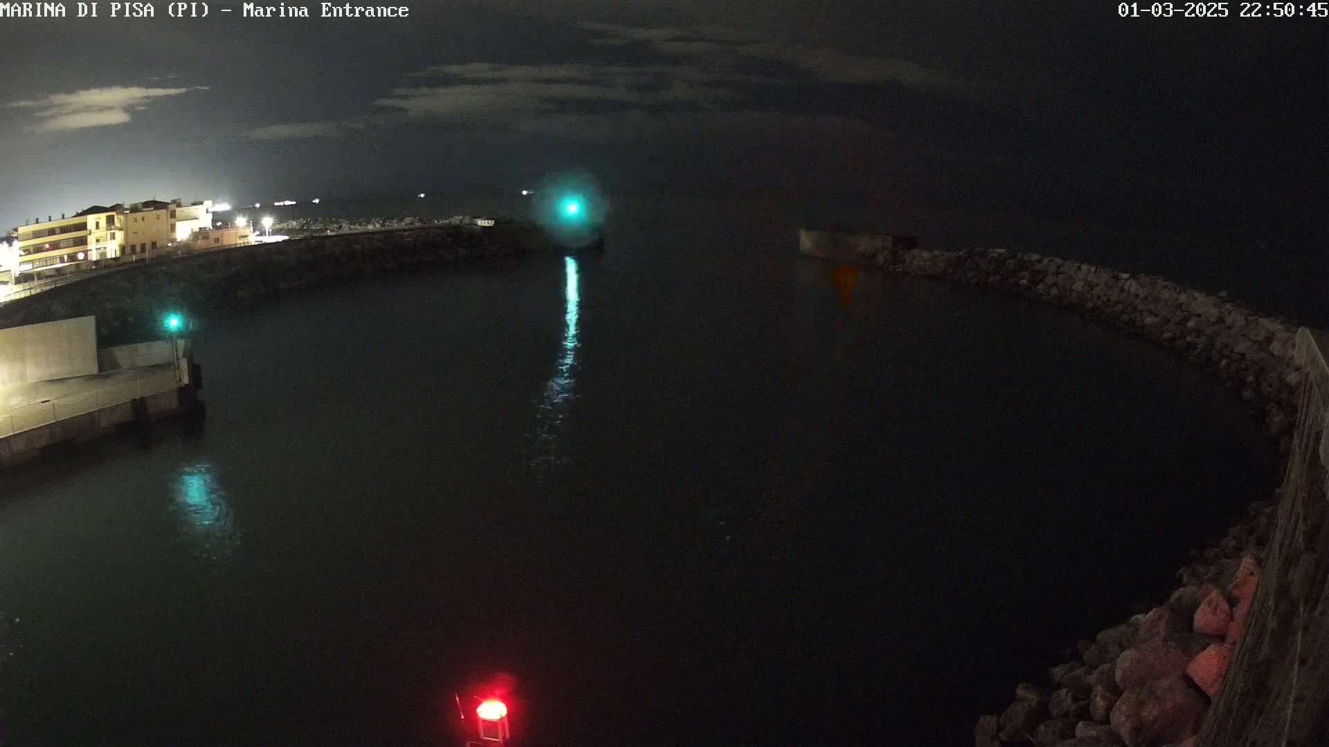 A nighttime view of a harbor entrance with calm water, illuminated by lights on the docks and a faint glow in the distance under a partly cloudy sky.