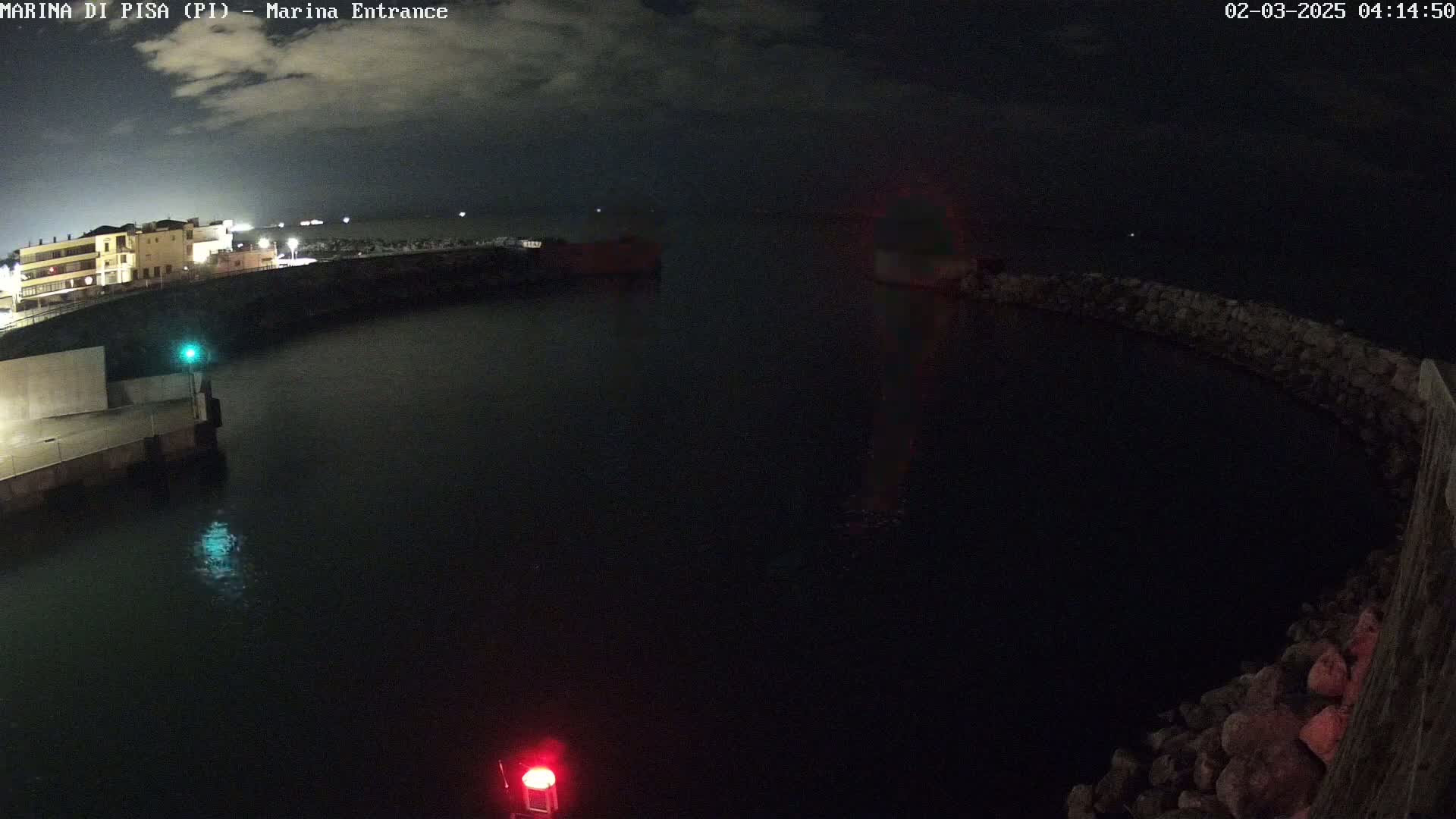 A nighttime view of a dark harbor with a building and some lights visible on the left, under a mostly cloudy sky.