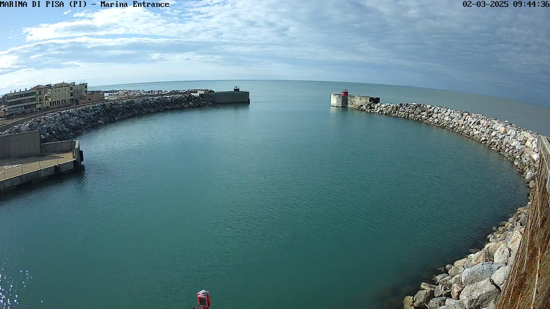 A harbor entrance with calm, teal water is bordered by rock walls and a building is visible in the background under a partly cloudy sky.