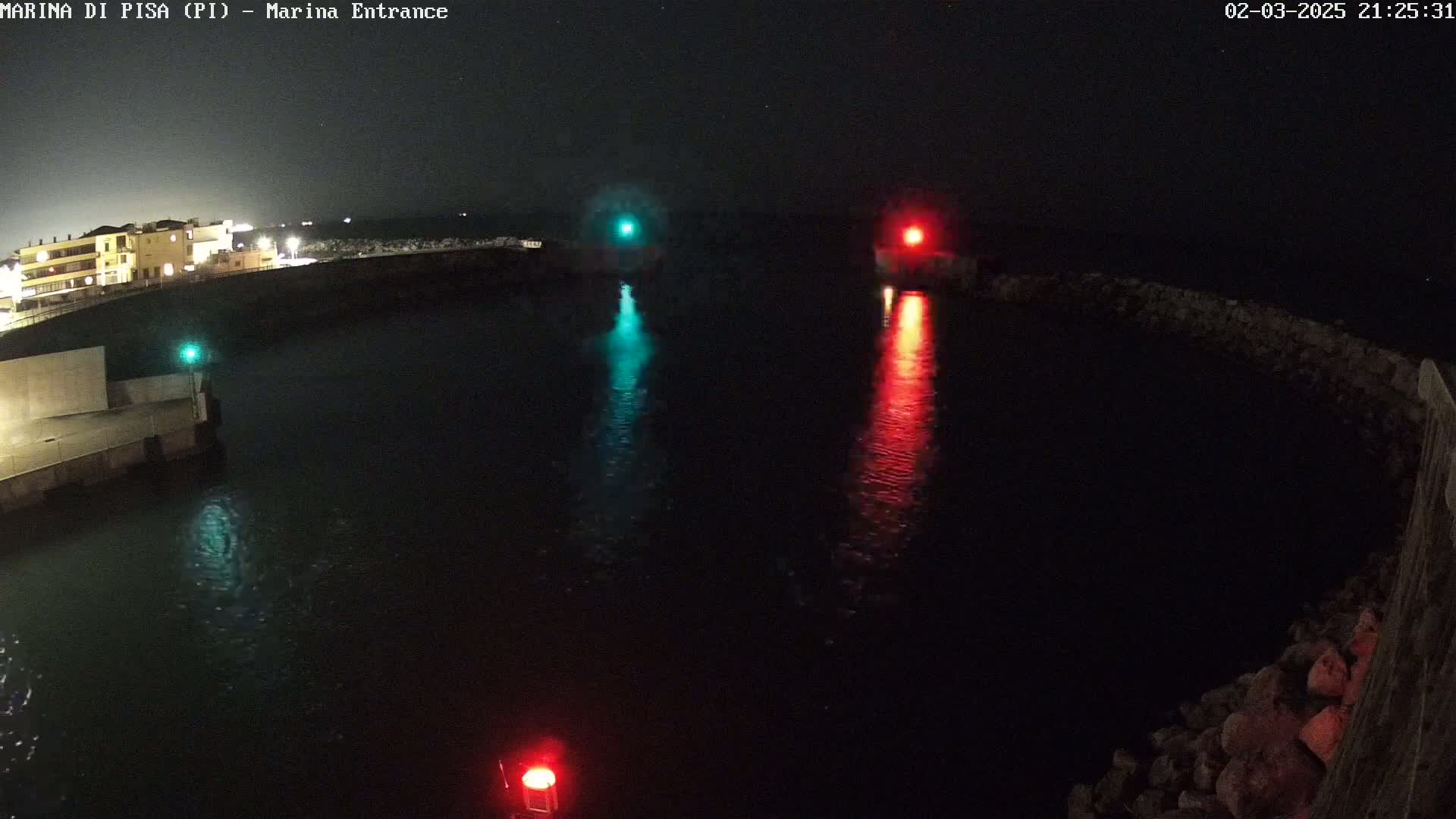A nighttime view of a harbor entrance with calm water reflecting green and red navigation lights.