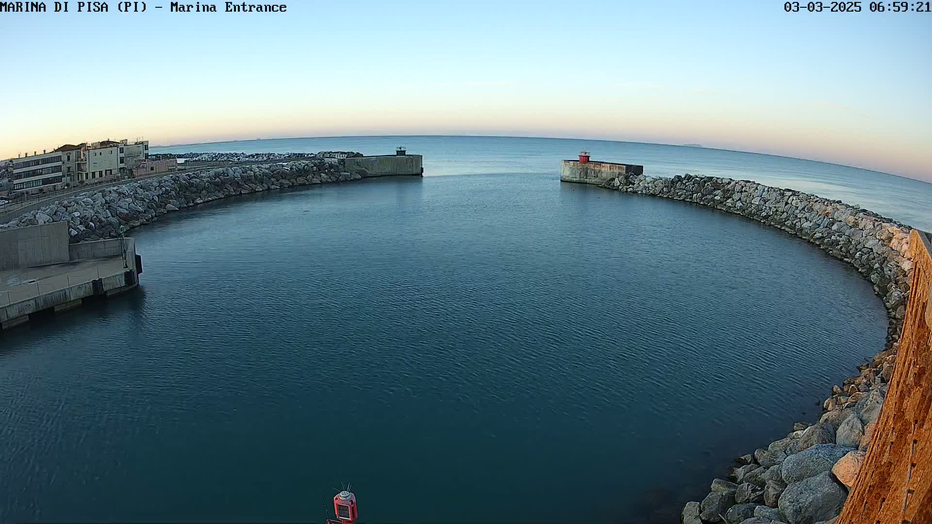 A calm, blue harbor is enclosed by a curved rock wall under a clear, light-blue sky.