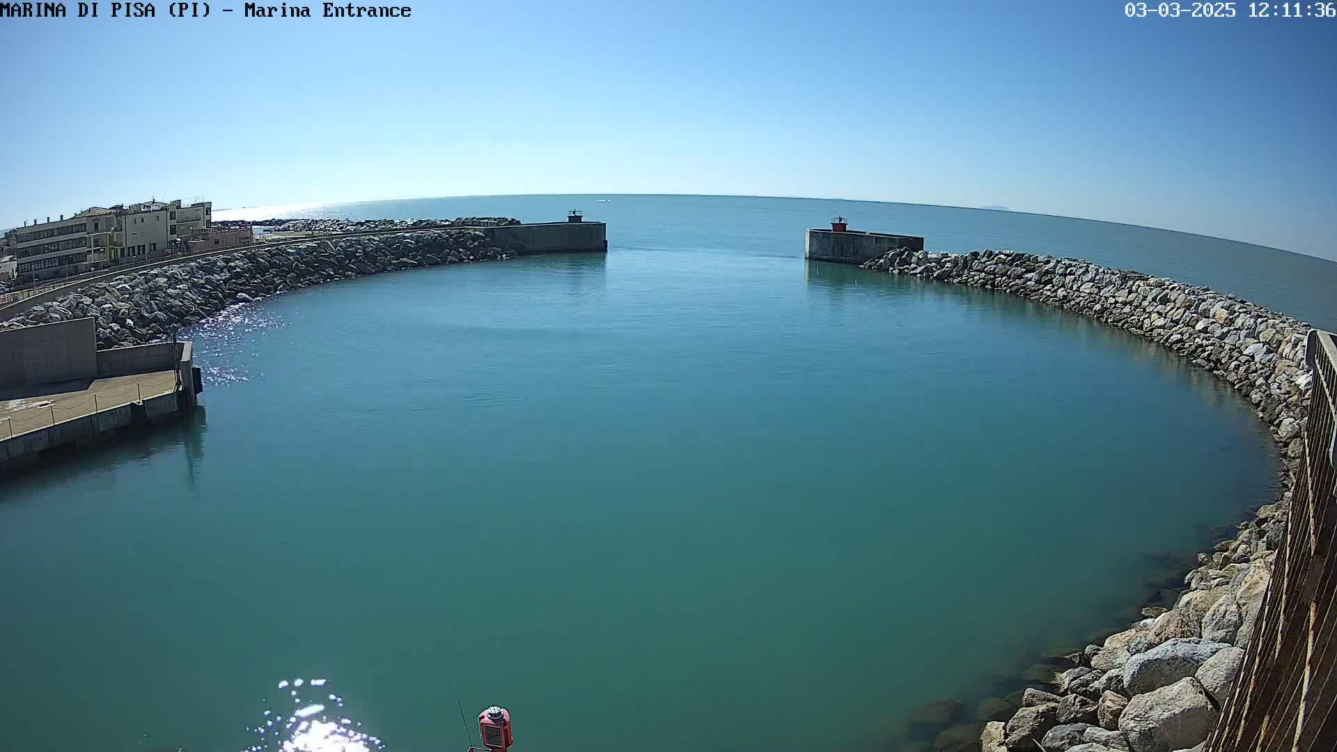 A harbor entrance with calm, teal water is partially enclosed by two rock walls extending from the shore under a clear blue sky.