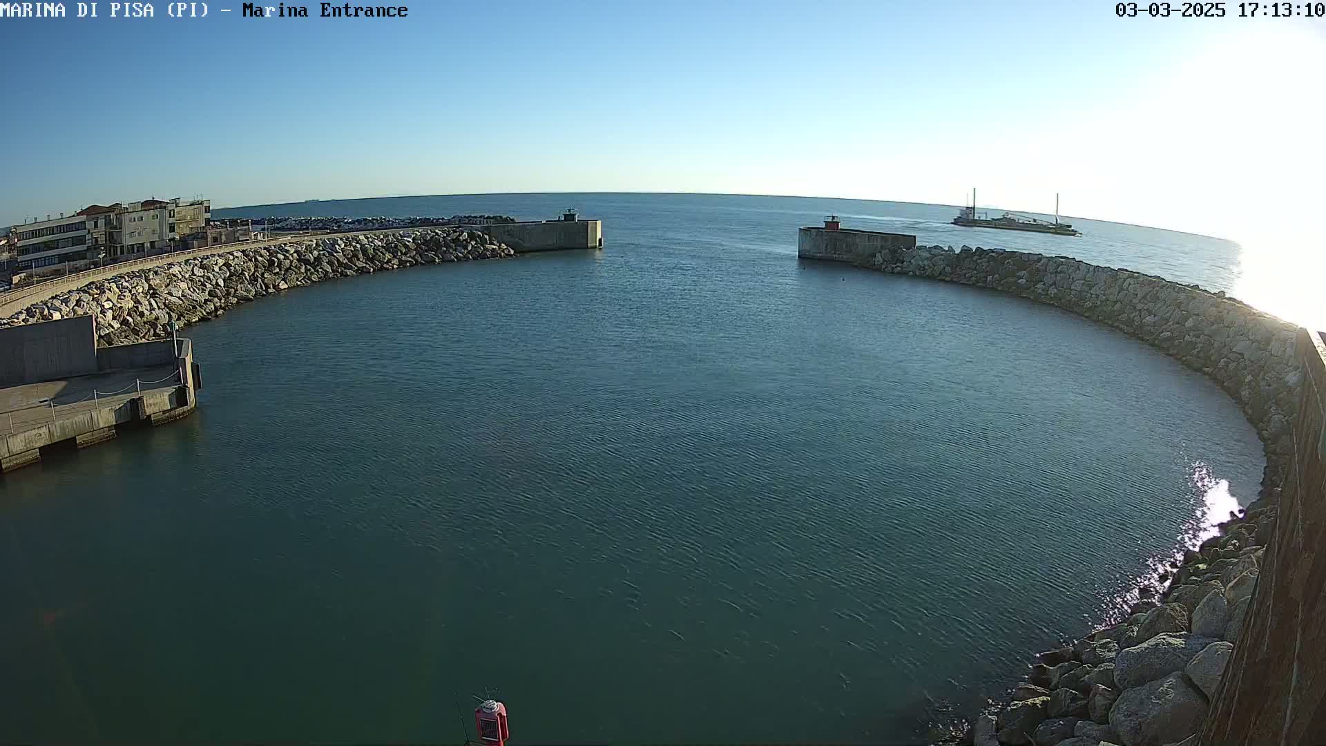 A harbor entrance with calm, blue water is enclosed by two curved rock walls, with a vessel visible in the distance under a clear sky.