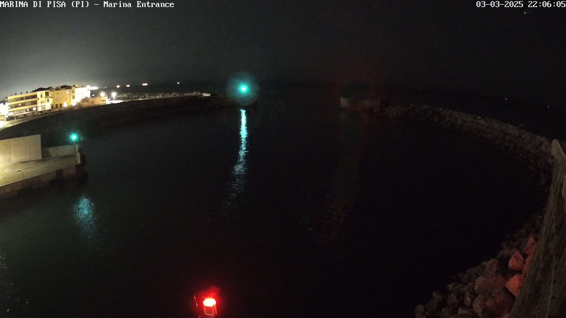 A nighttime view of a harbor entrance with calm water reflecting a green light, illuminated buildings on the shore, and a rocky seawall.