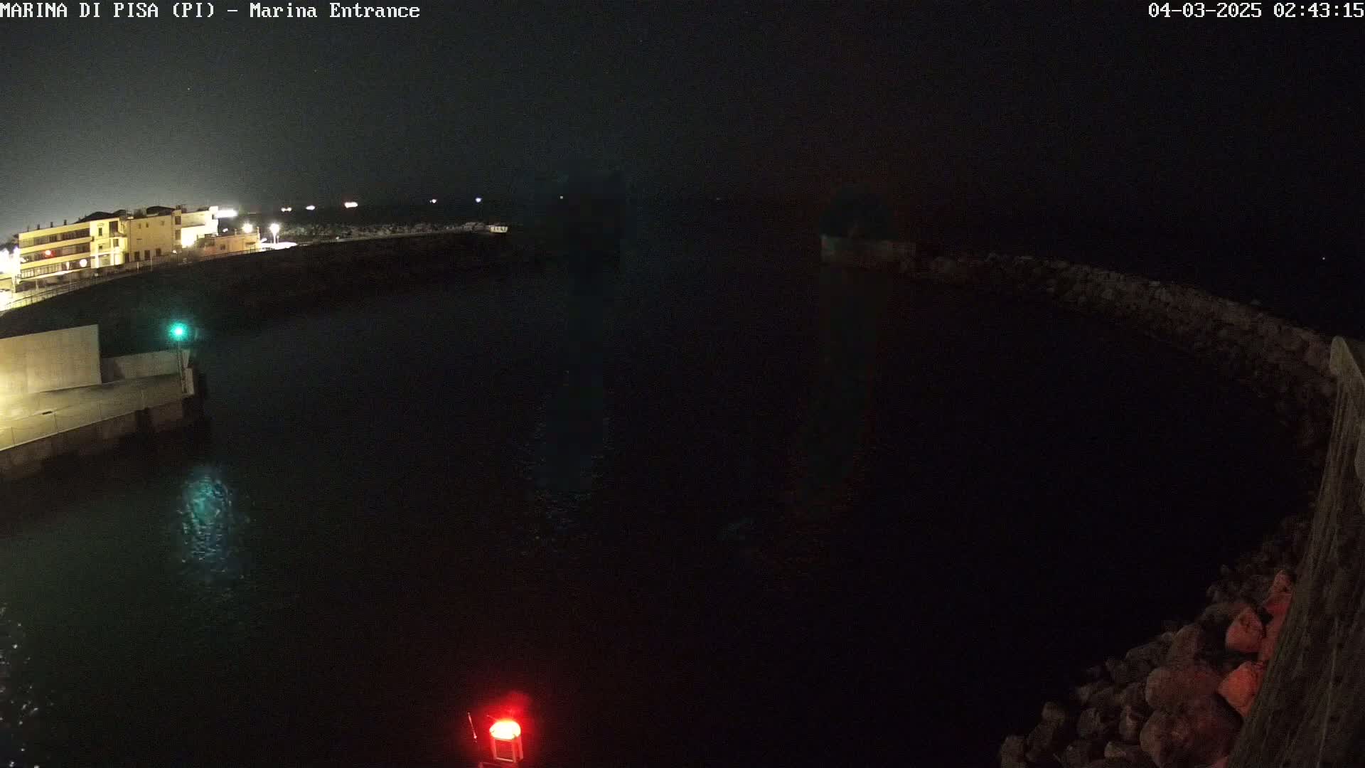 A dark nighttime view of a harbor entrance with calm water, showing a building with lights on in the distance.