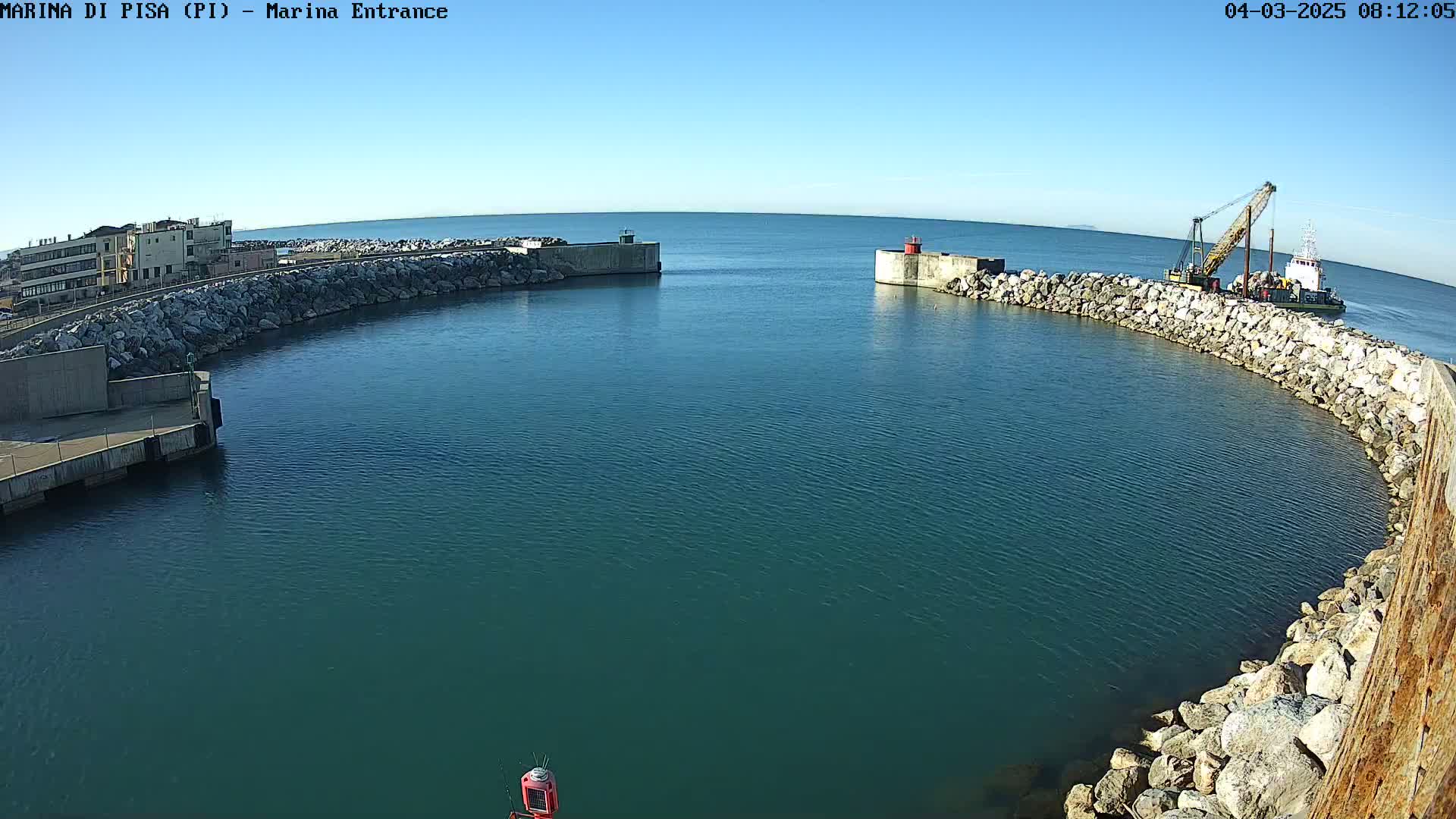 A harbor entrance with calm, clear blue water, surrounded by rock walls, and a ship working near one of the walls under a clear blue sky.
