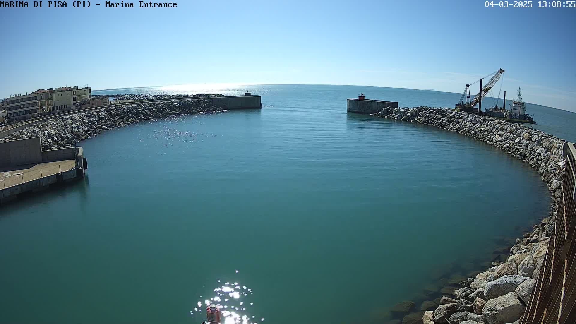 A harbor entrance with teal water under a clear, sunny sky shows a crane ship near a rocky wall.
