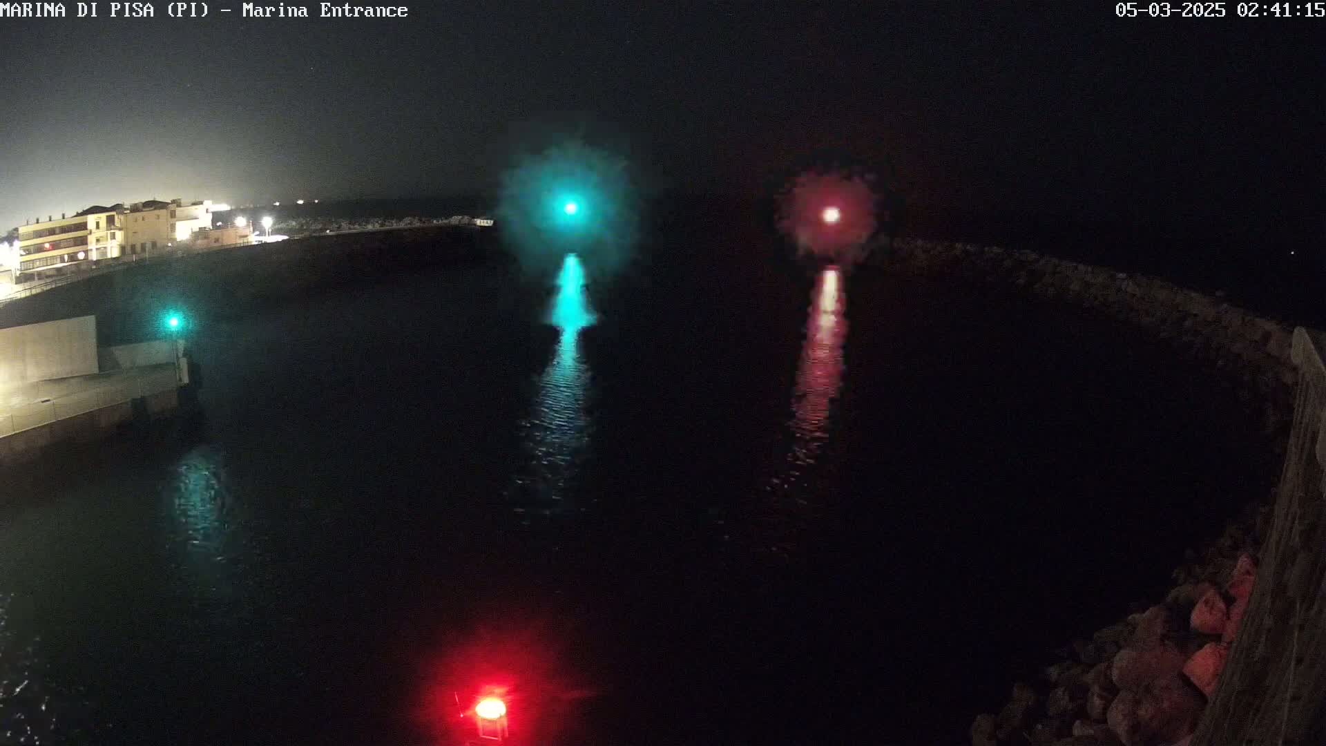 At night, two bursts of colored light, one teal and one red, illuminate a dark body of water near a stone wall and buildings.