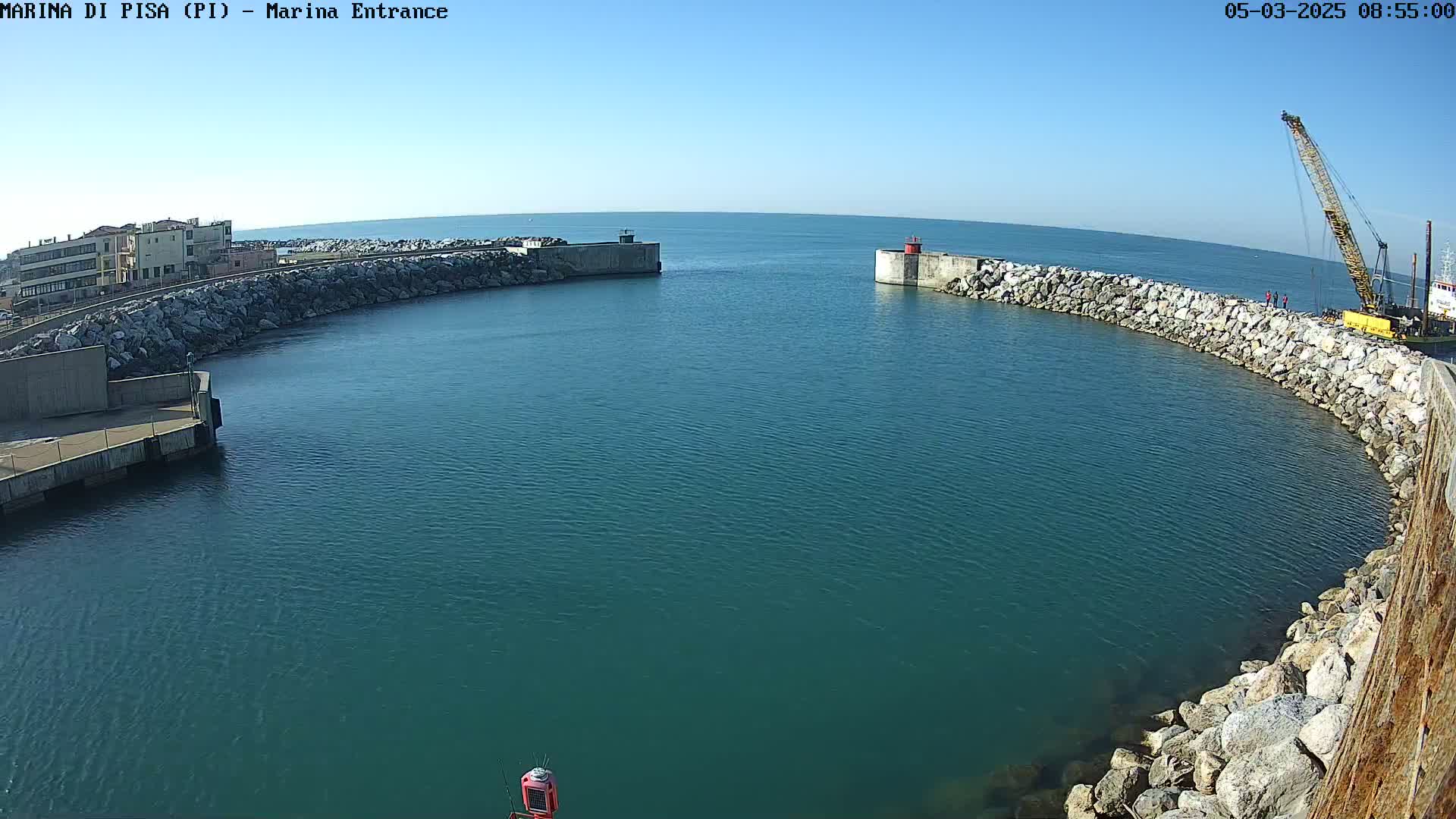 A harbor entrance with calm, teal water is partially enclosed by rock walls, with a building and crane visible on the shore under a clear blue sky.