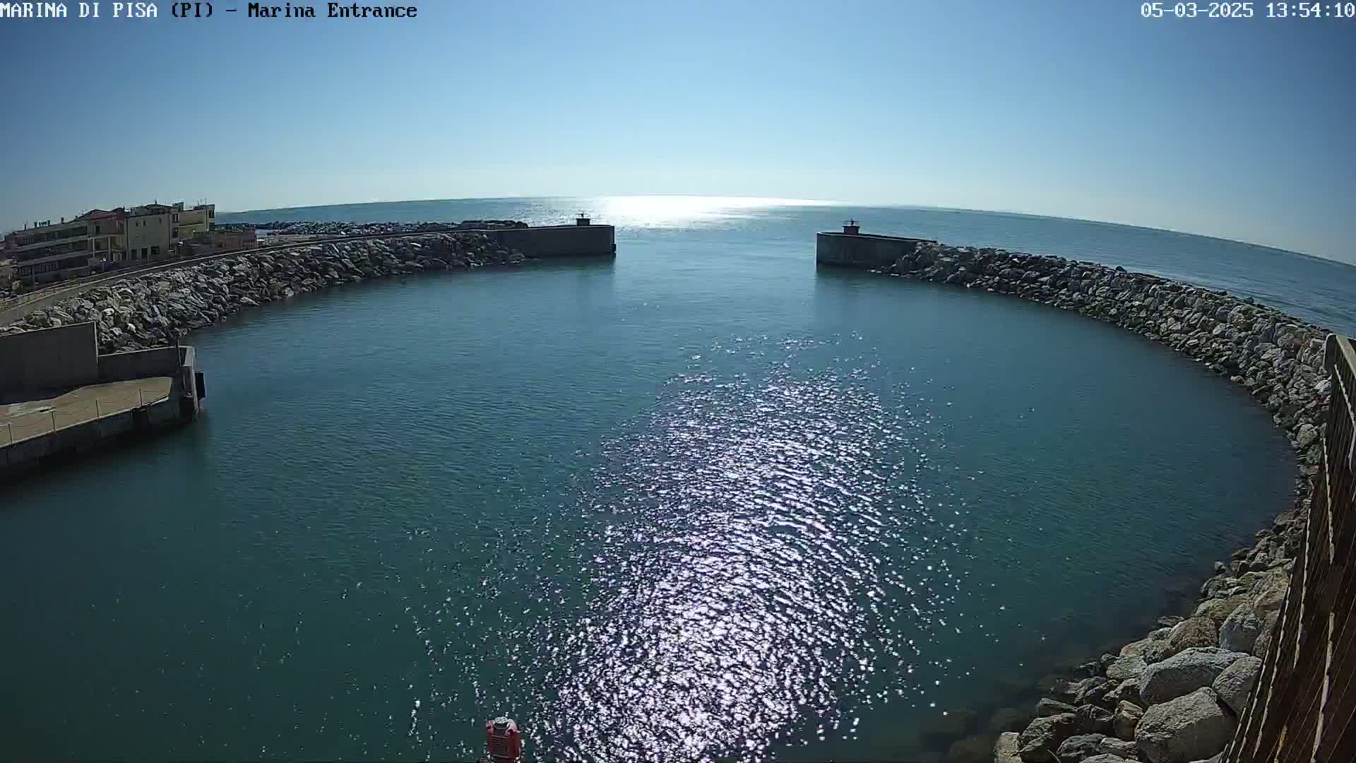 A harbor entrance with calm, sunlit water is partially enclosed by rock walls, extending to buildings on the shore under a clear blue sky.