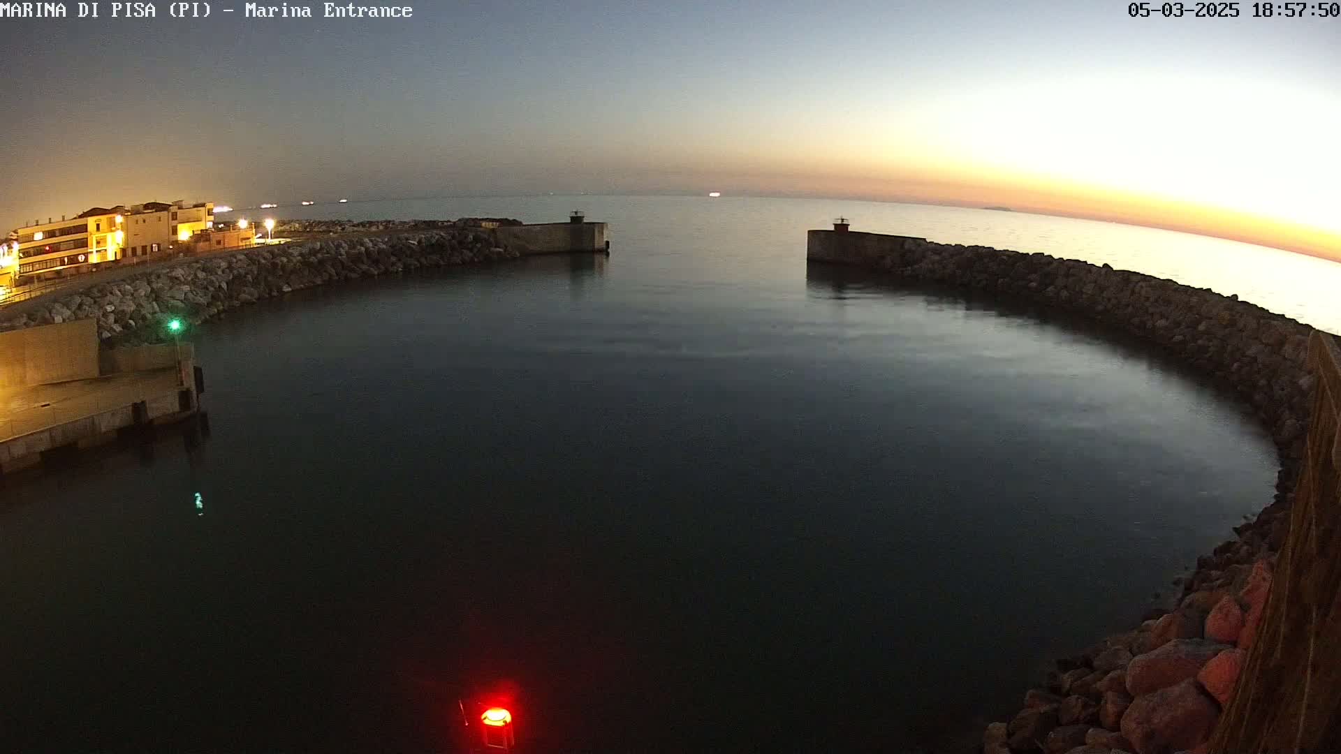 A calm, dark body of water is enclosed by a curved stone wall at dusk, with a building and lights visible in the background.