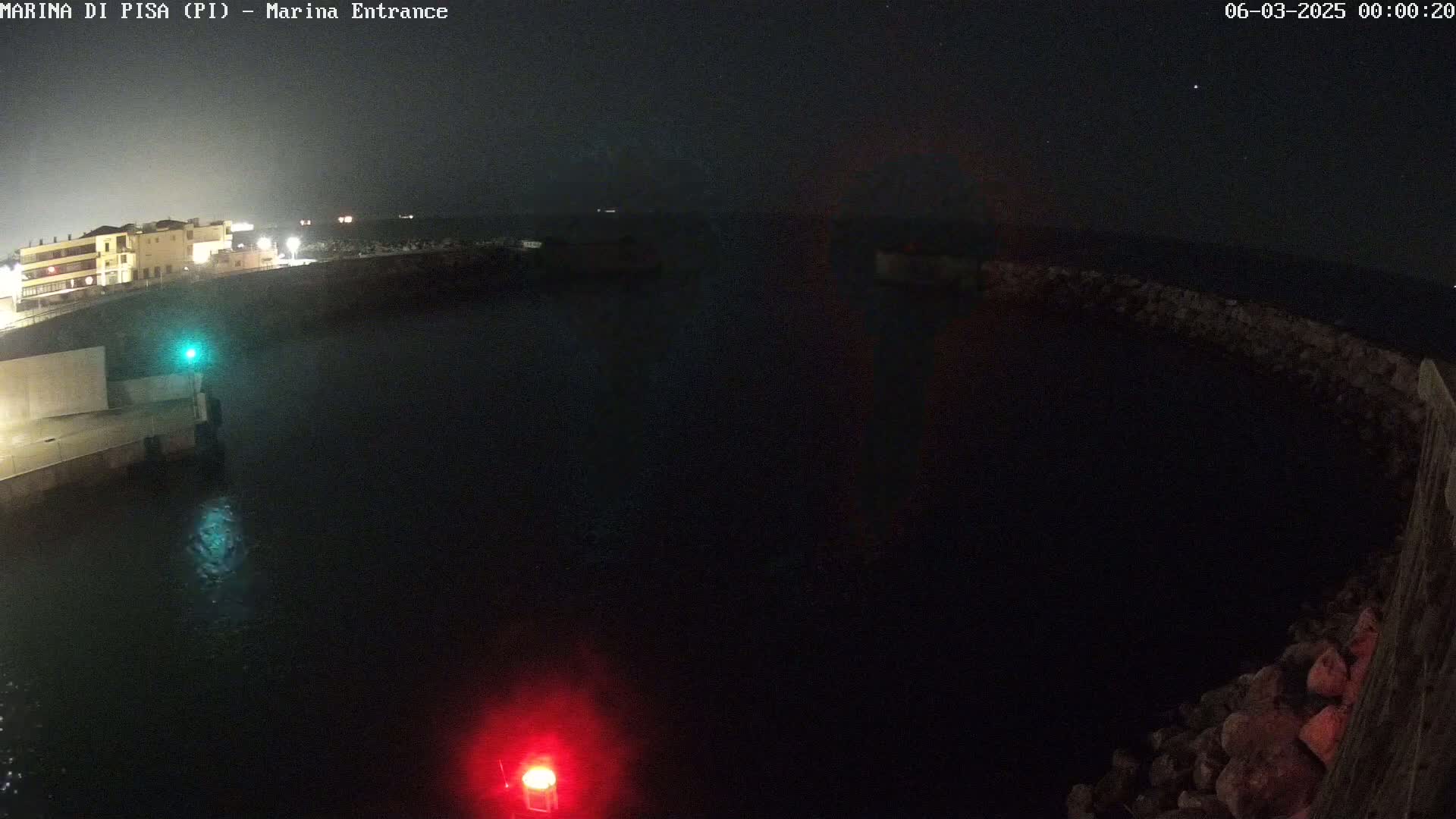 A nighttime, dark view of a marina entrance with calm water, showing a building and stone walls on either side of the waterway.