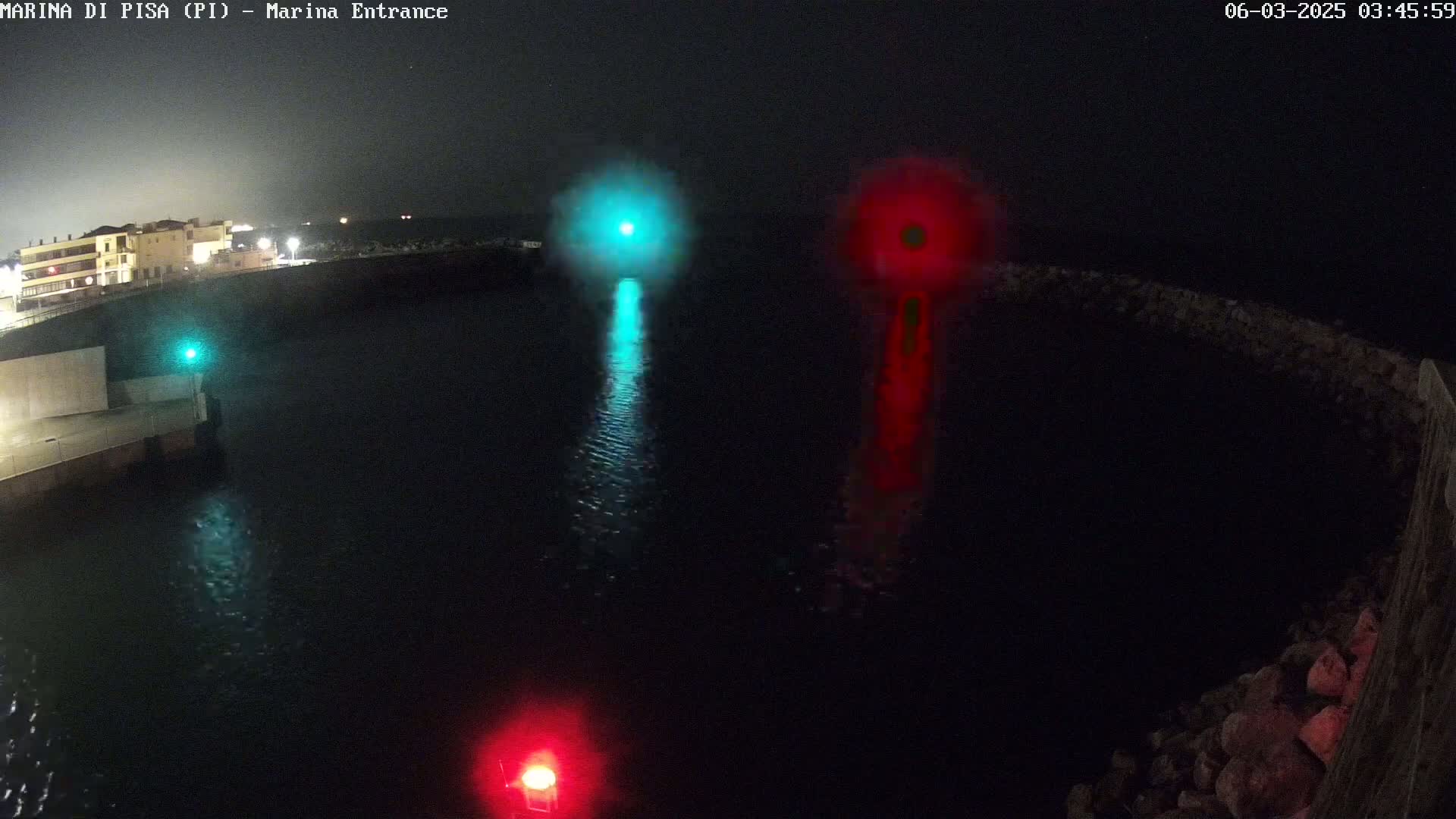 At night, a dark harbor is illuminated by a green and a red light, reflecting on the water's surface near a stone wall and a building in the distance.