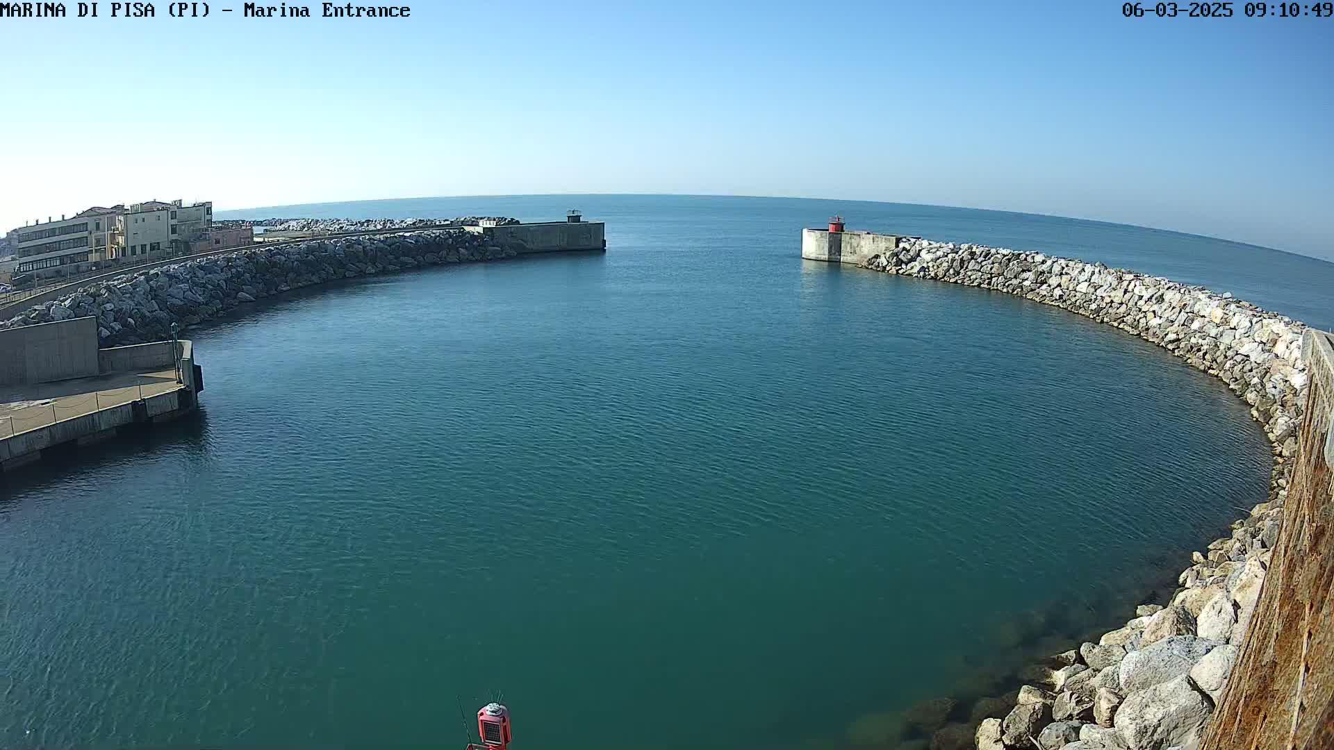 A harbor entrance with calm, teal water is surrounded by rock walls and a building is visible in the background under a clear blue sky.