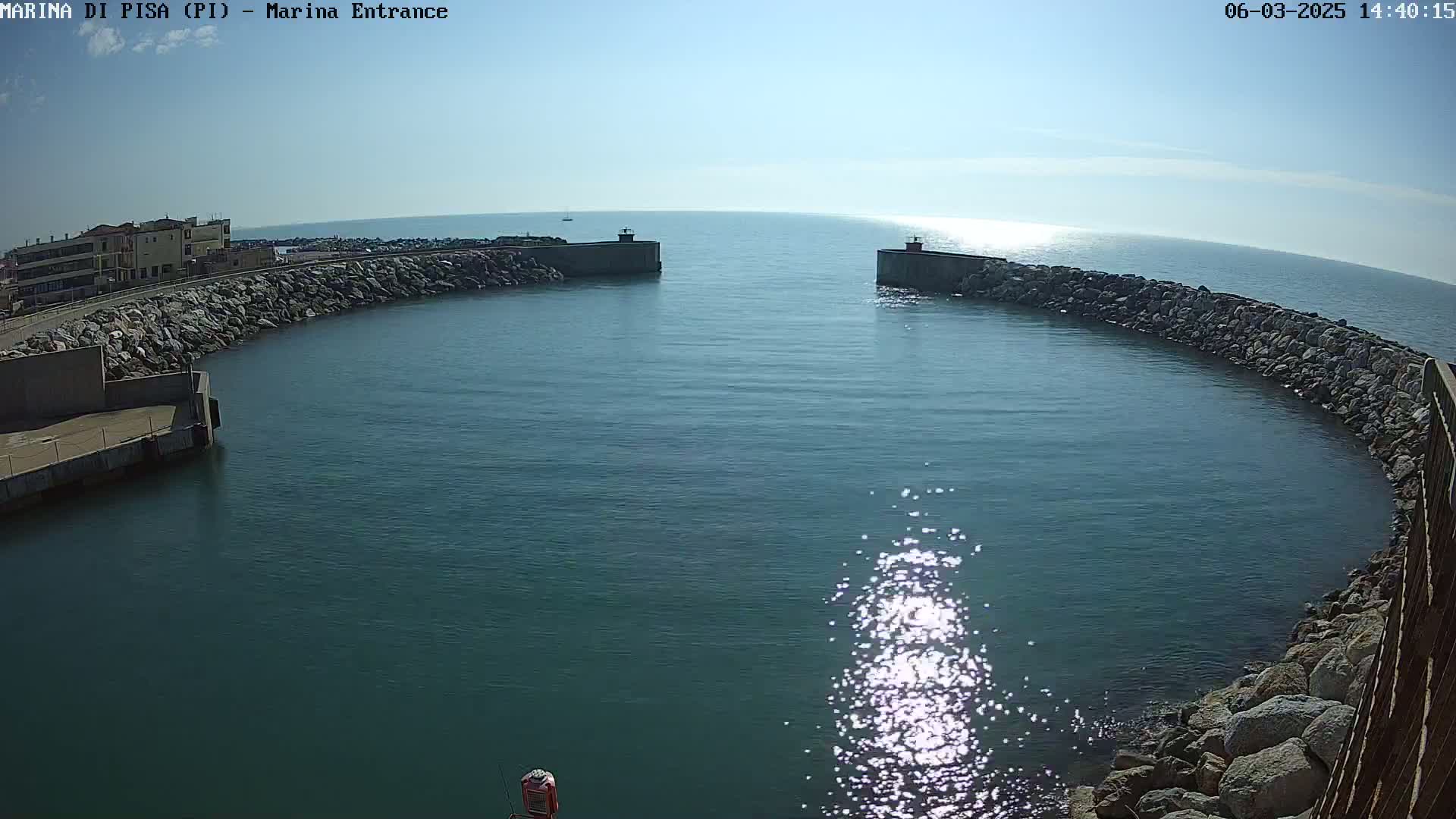 A harbor entrance with calm, sunlit water is partially enclosed by two rock-lined walls, under a clear sky.