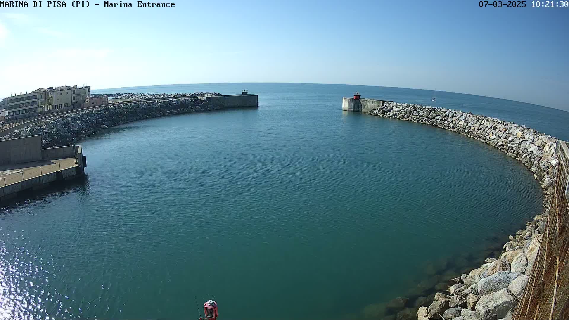 A marina entrance with calm, teal water is bordered by rock walls under a clear blue sky.