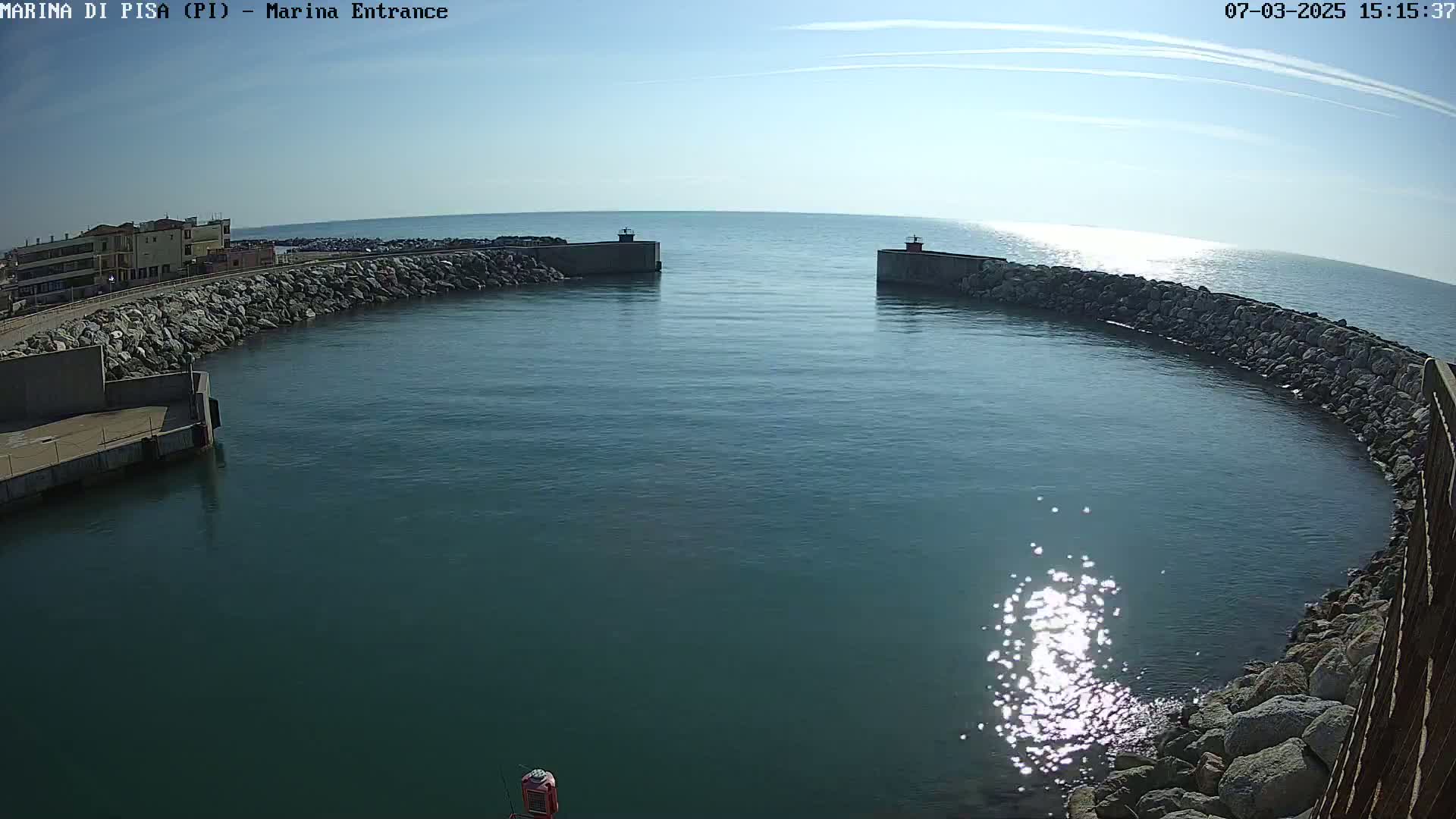 A calm, sunny day reveals a harbor entrance, defined by curving rock walls and a tranquil expanse of dark blue water reflecting sunlight.