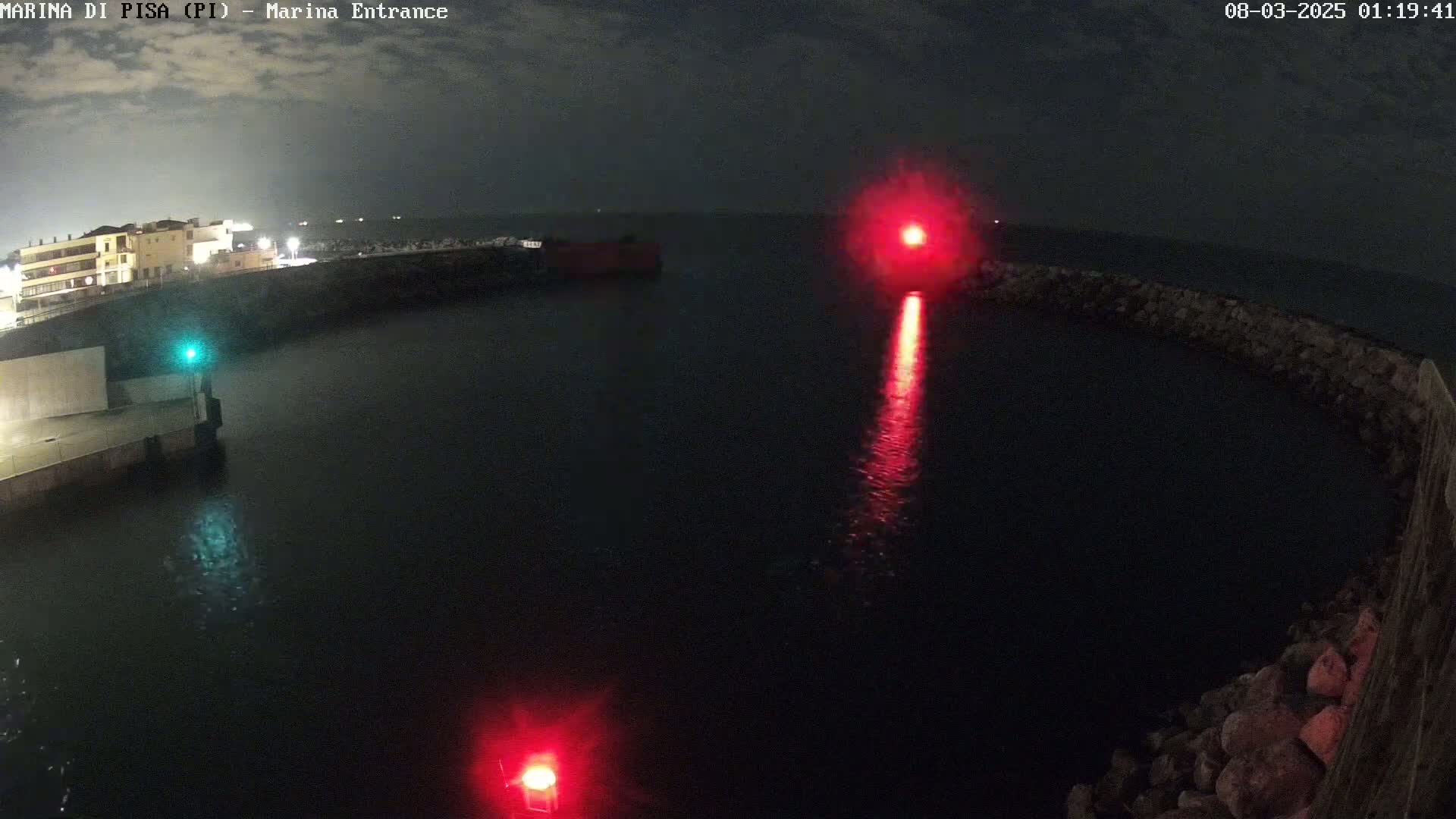 A nighttime aerial view of a harbor entrance with two red navigation lights visible on the water, under a partly cloudy sky.