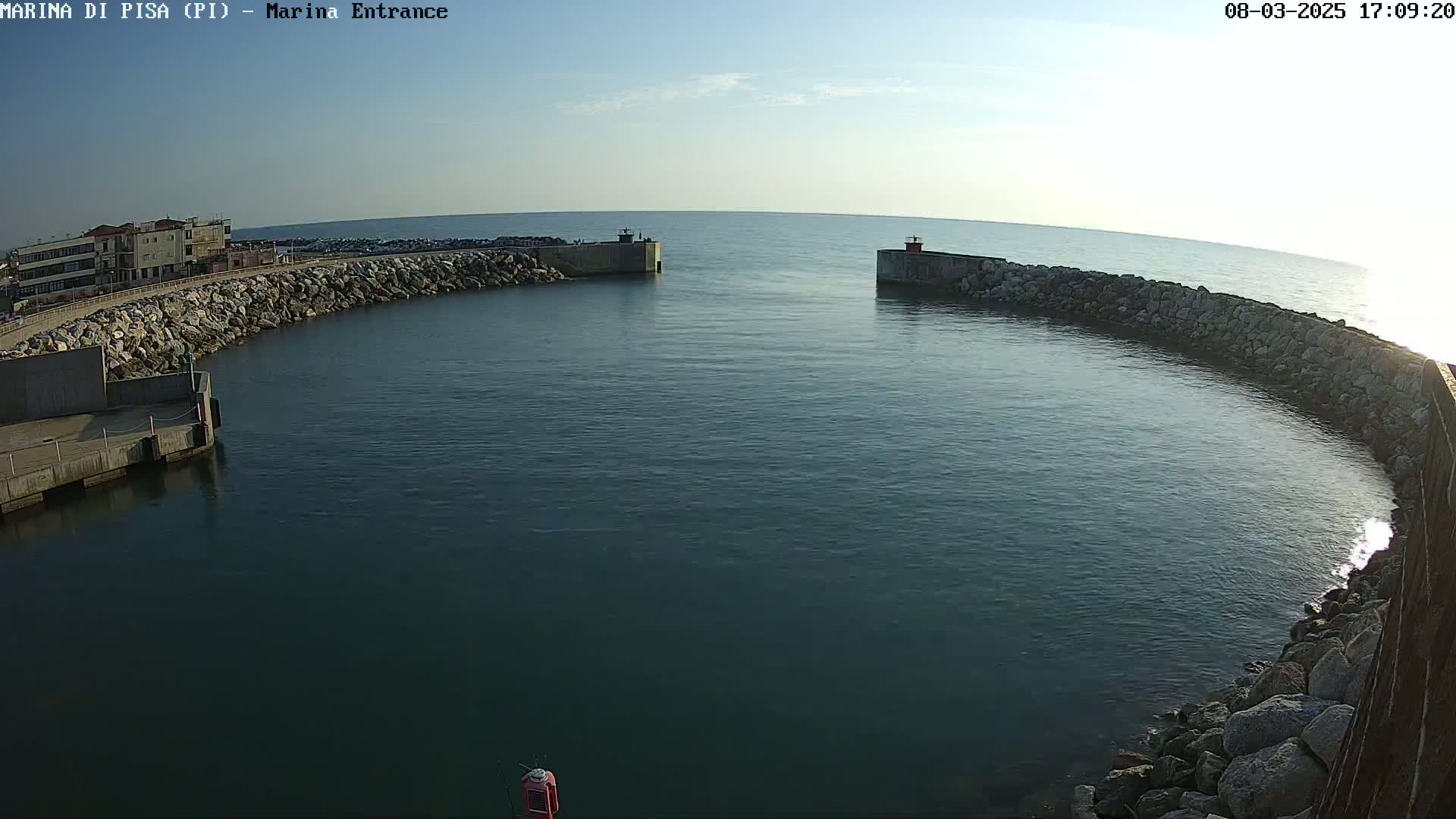 A harbor entrance with calm water under a clear, bright sky is framed by rocky breakwaters.