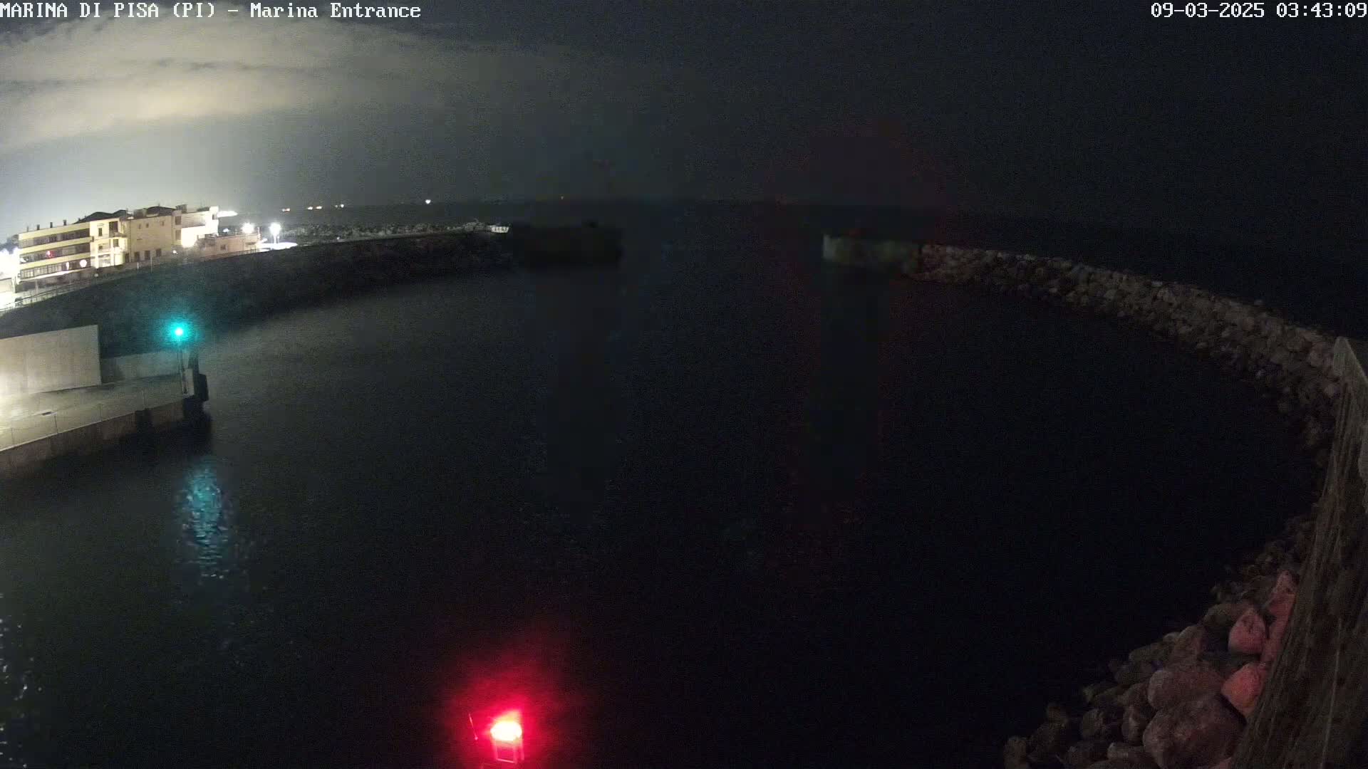 A nighttime view of a dark harbor with a building in the background, calm water, and a rocky breakwater.