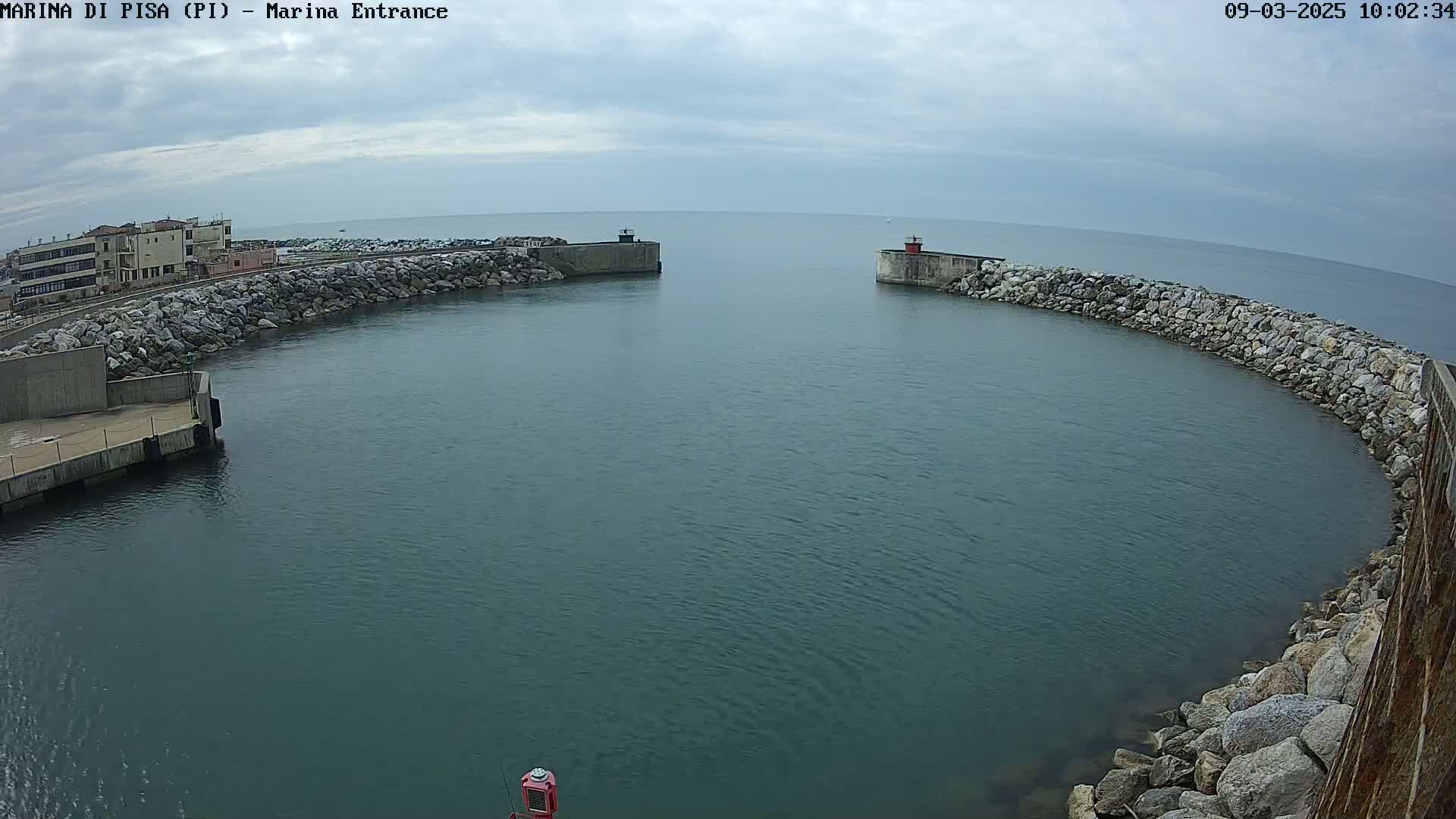 A calm, overcast day shows a harbor entrance with two rock breakwaters curving towards a body of dark-blue water.
