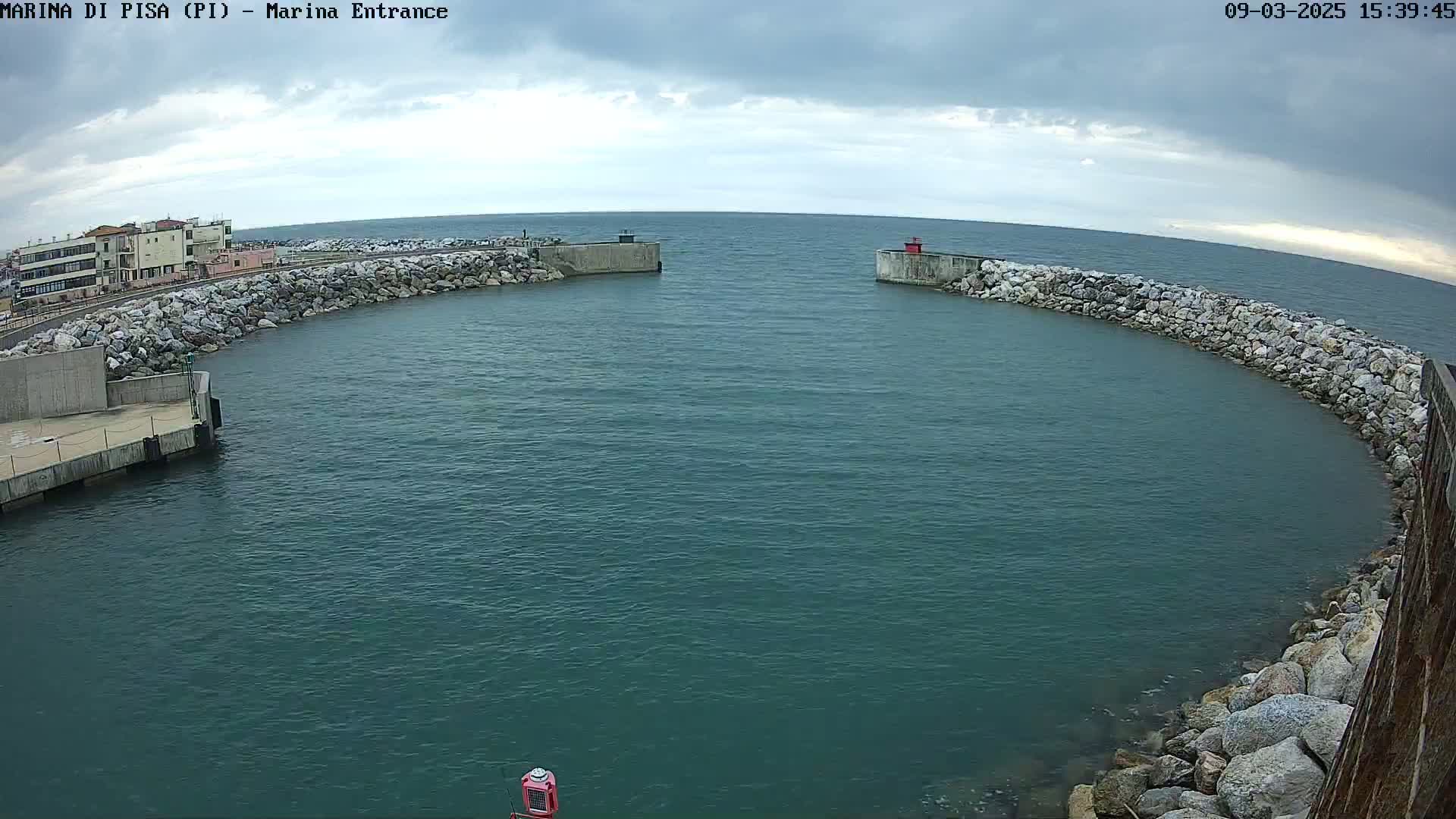A harbor entrance with calm, dark-blue water is partially enclosed by a curved rock wall under a cloudy sky.