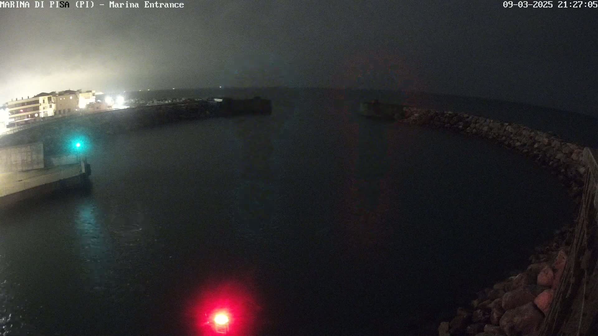 A nighttime view of a dark harbor entrance with a building faintly visible in the distance under an overcast sky.
