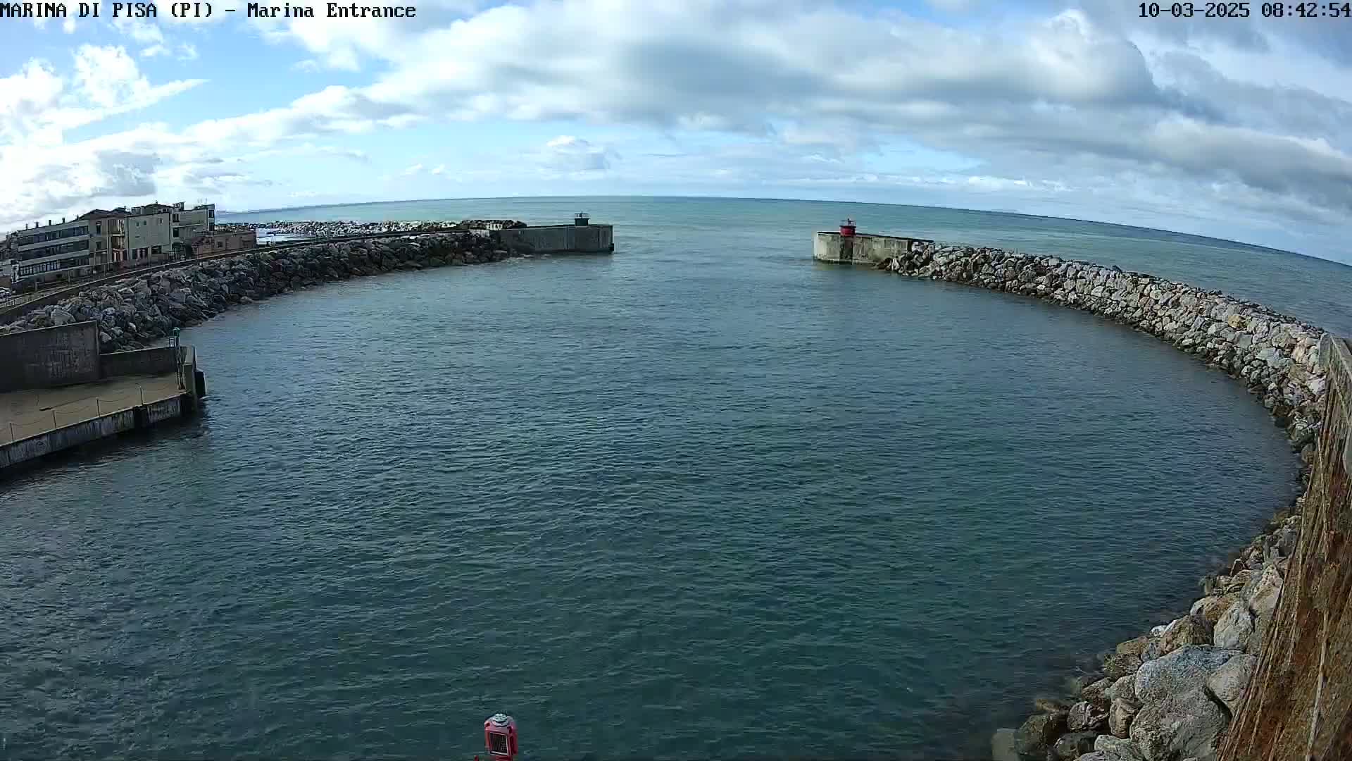 A partly cloudy day reveals a harbor entrance framed by stone walls, with calm water and buildings visible in the background.