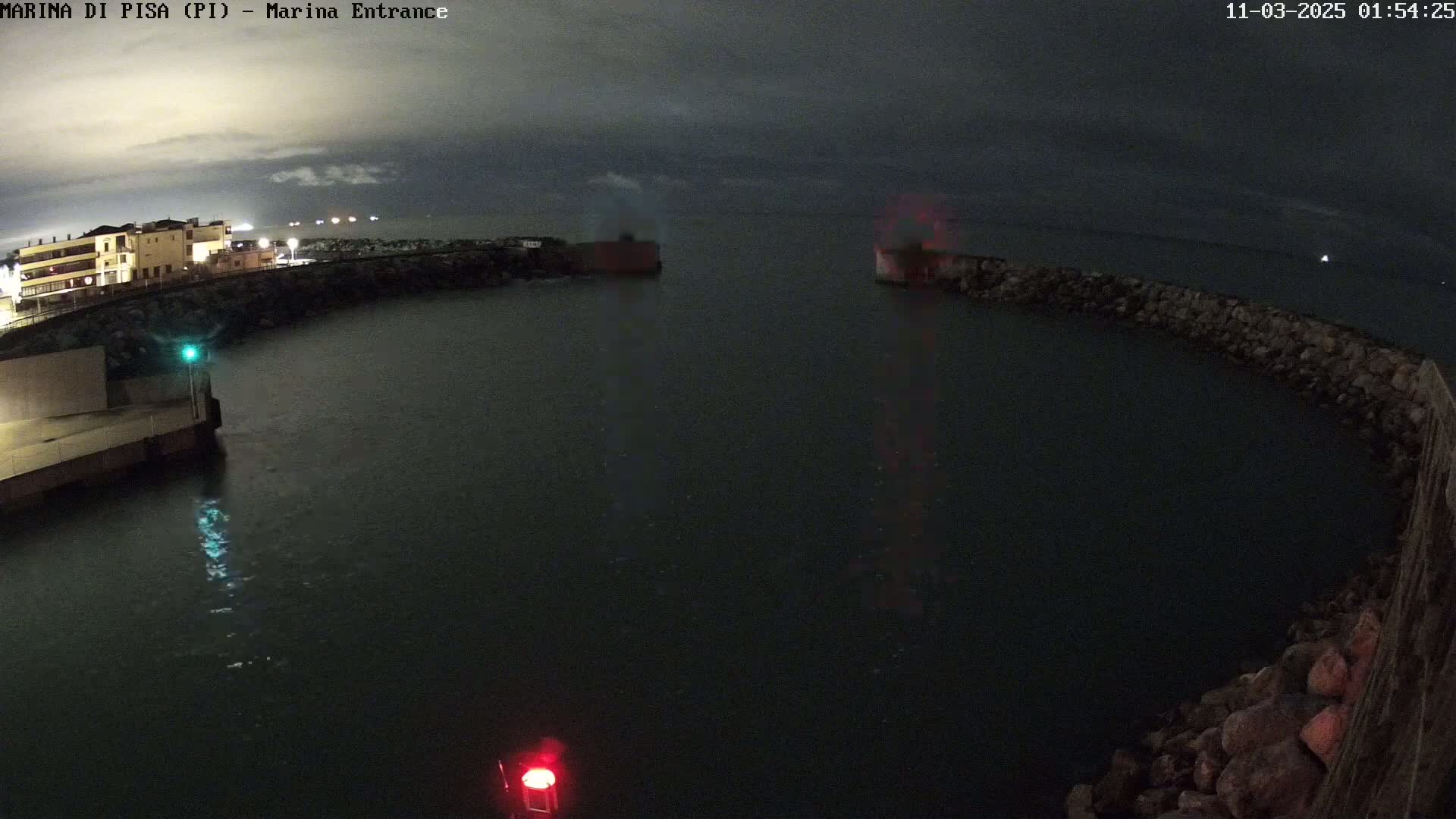 A nighttime view of a calm harbor entrance with buildings in the background under a dark, cloudy sky.