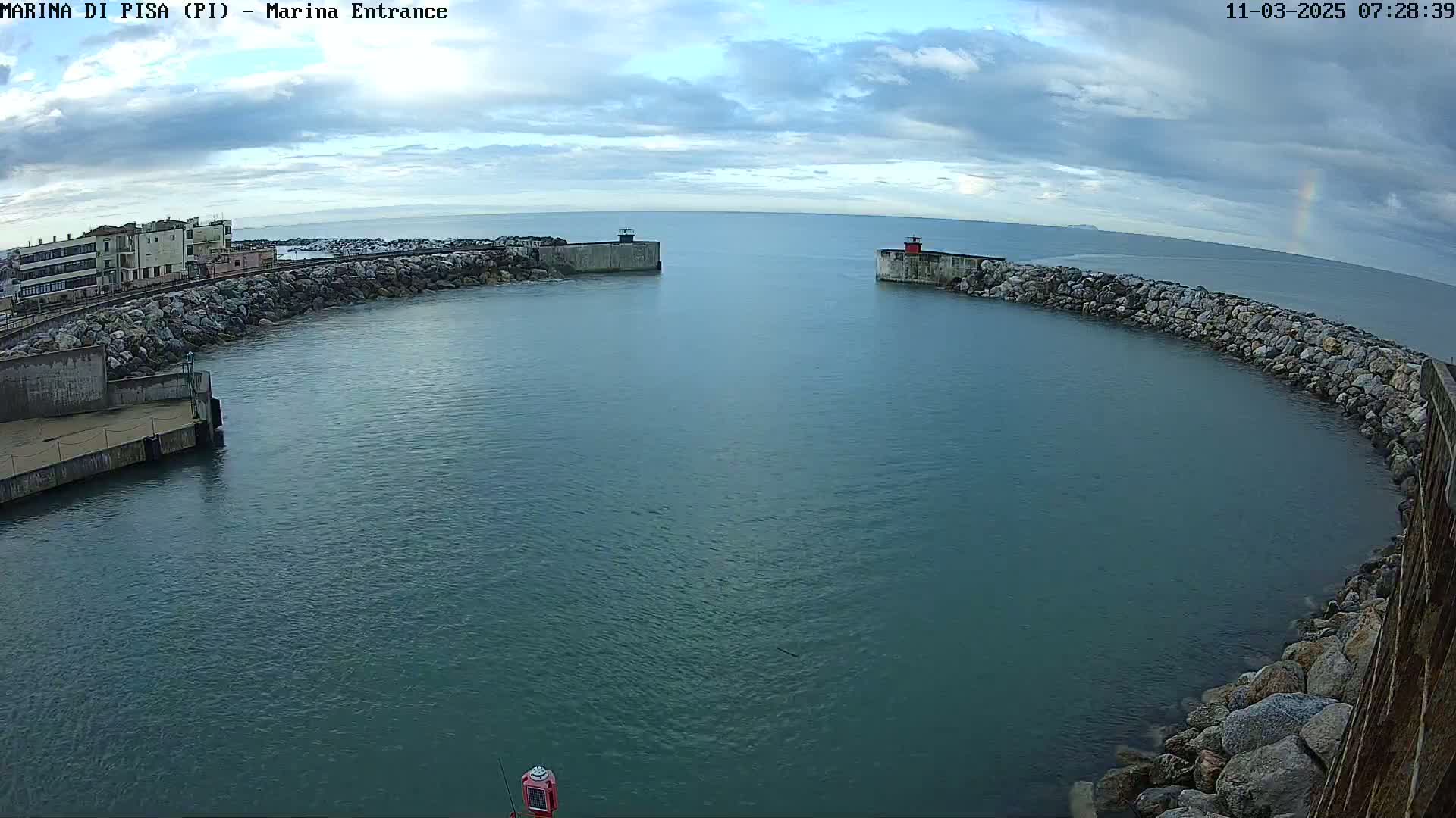 A harbor entrance with calm, teal water under a partly cloudy sky, a faint rainbow visible in the distance.