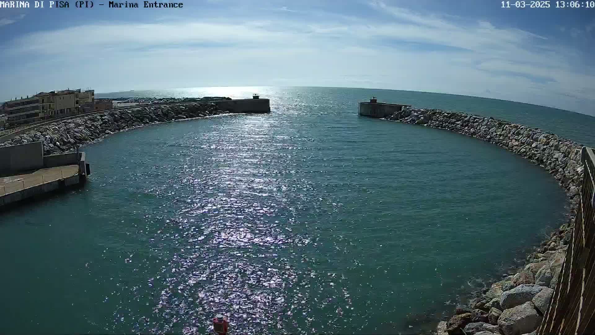 A harbor entrance with calm, sunlit water is nestled between two rocky breakwaters on a partly sunny day.