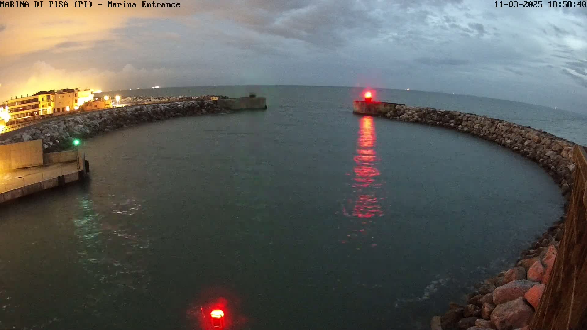 A harbor entrance at dusk under a cloudy sky, showing calm water partially enclosed by a stone wall with a red light on one side.