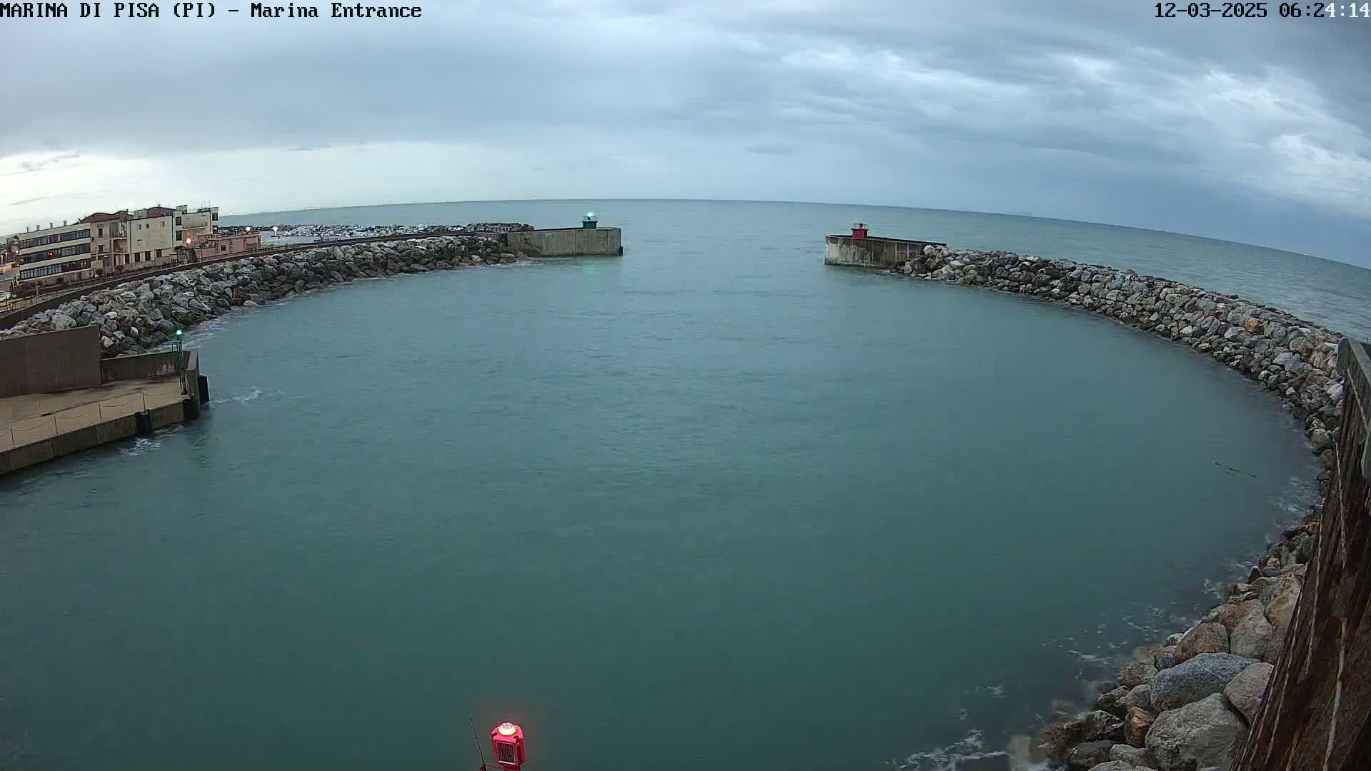 A calm, overcast day reveals a harbor entrance marked by stone walls and small lighthouses, with buildings visible in the distance.