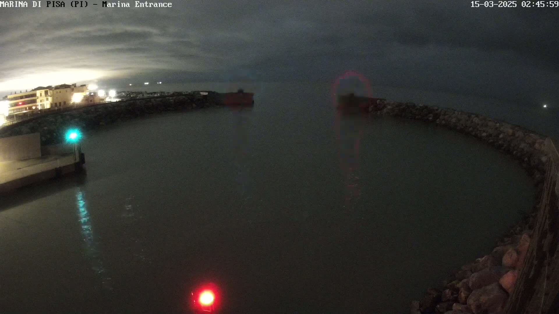 A nighttime view of a harbor entrance with calm water under a cloudy sky, showing a building with lights on and two vessels in the distance.