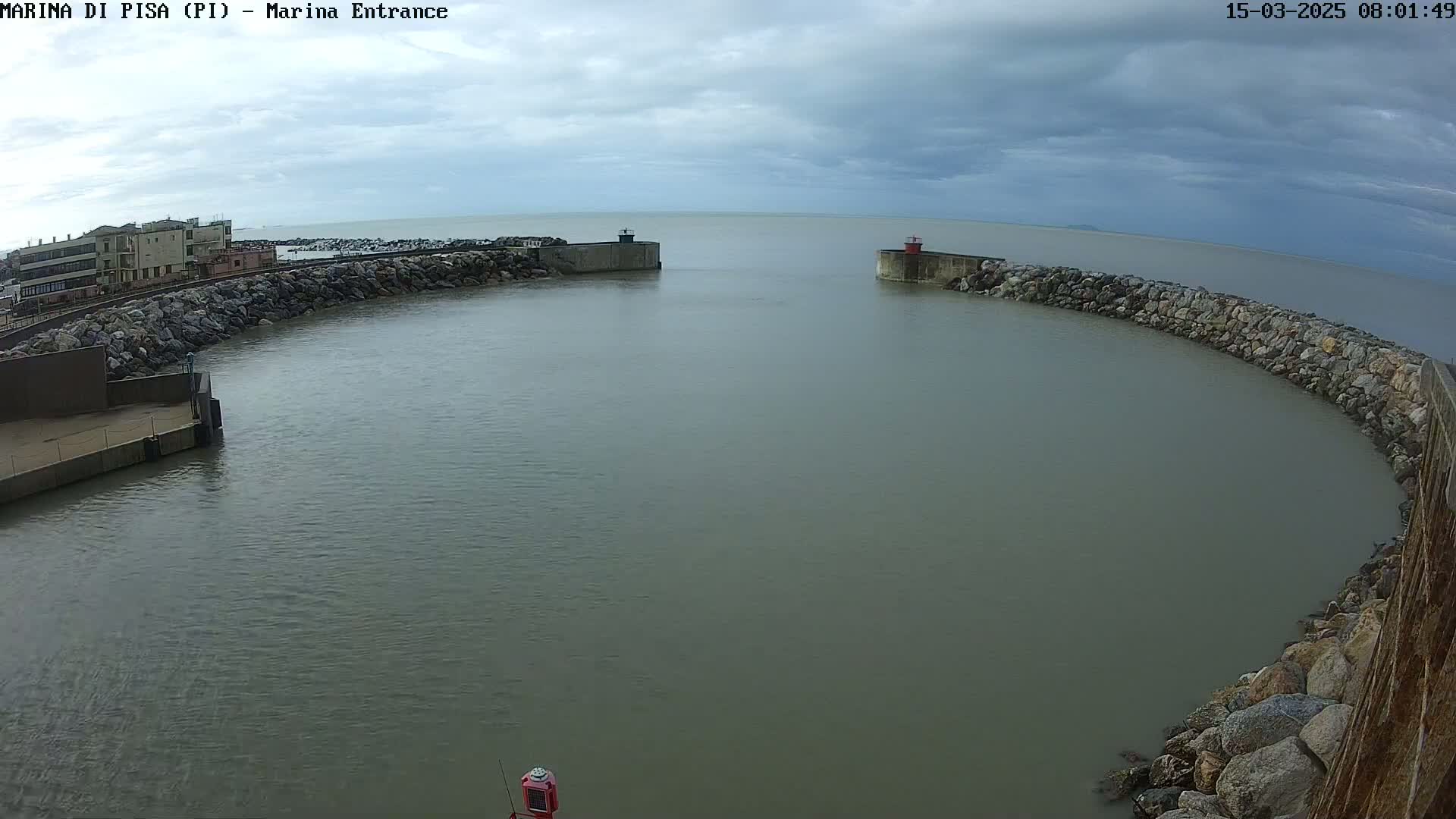 A harbor entrance with calm, gray water under a cloudy sky is bordered by rock walls and buildings.