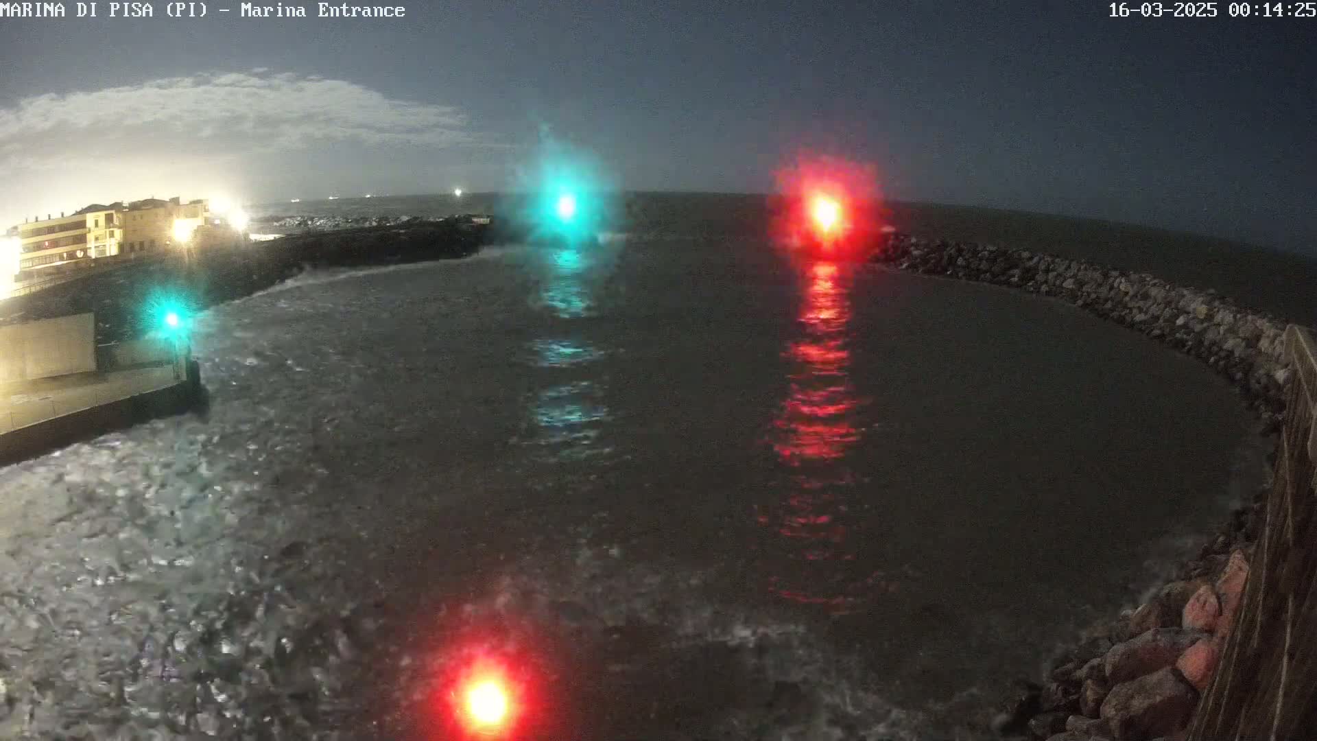 Under a dark, cloudy night sky, choppy water laps against a rocky shoreline near a building, illuminated by a green and a red light.