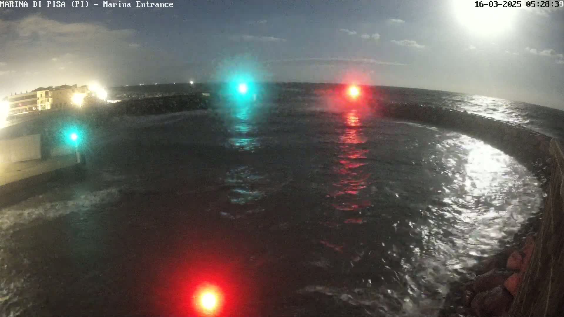 A nighttime view of choppy water with red and green lights on either side of a curved seawall.