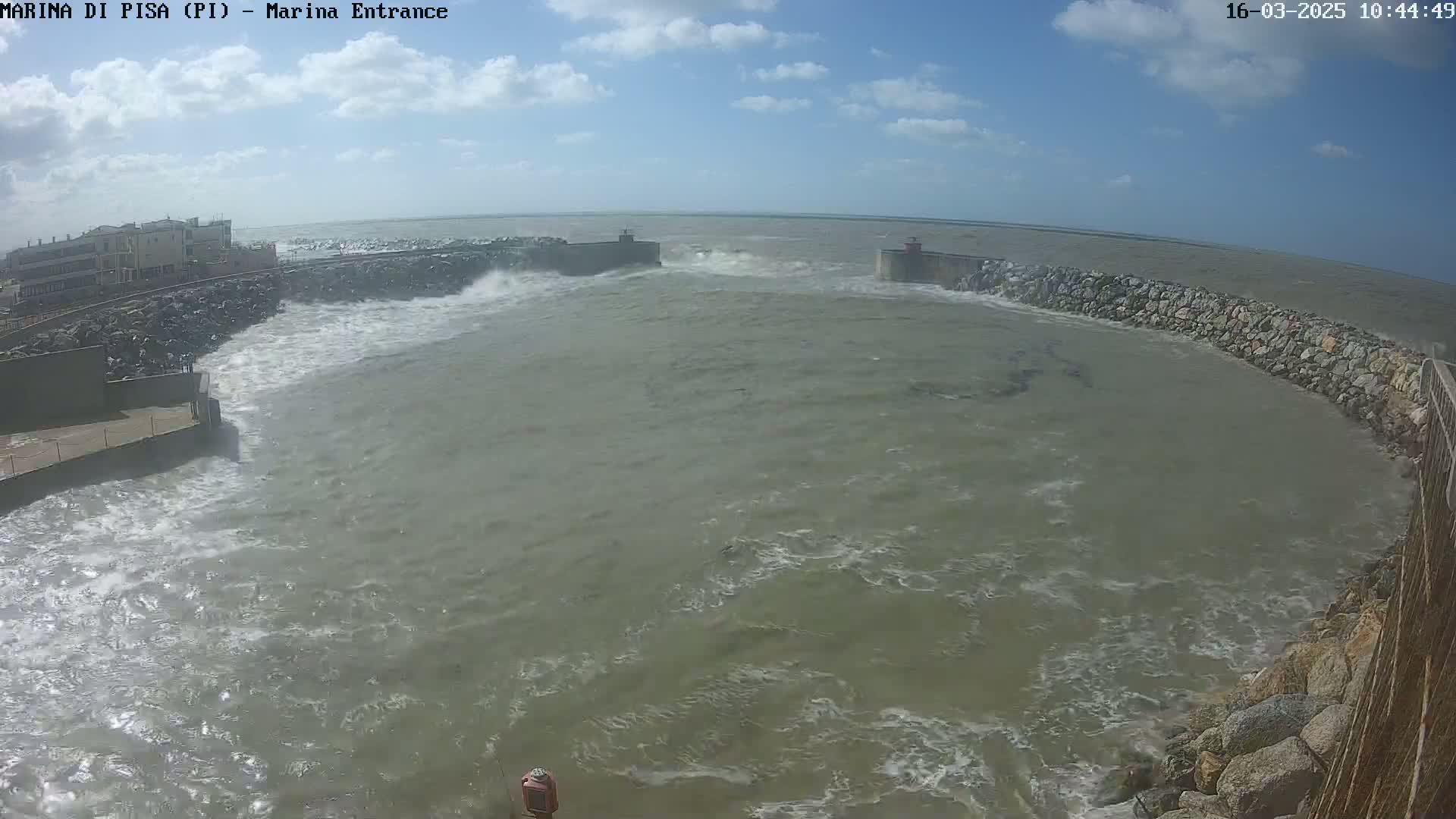Rough waves crash against a stone harbor wall under a partly cloudy sky.
