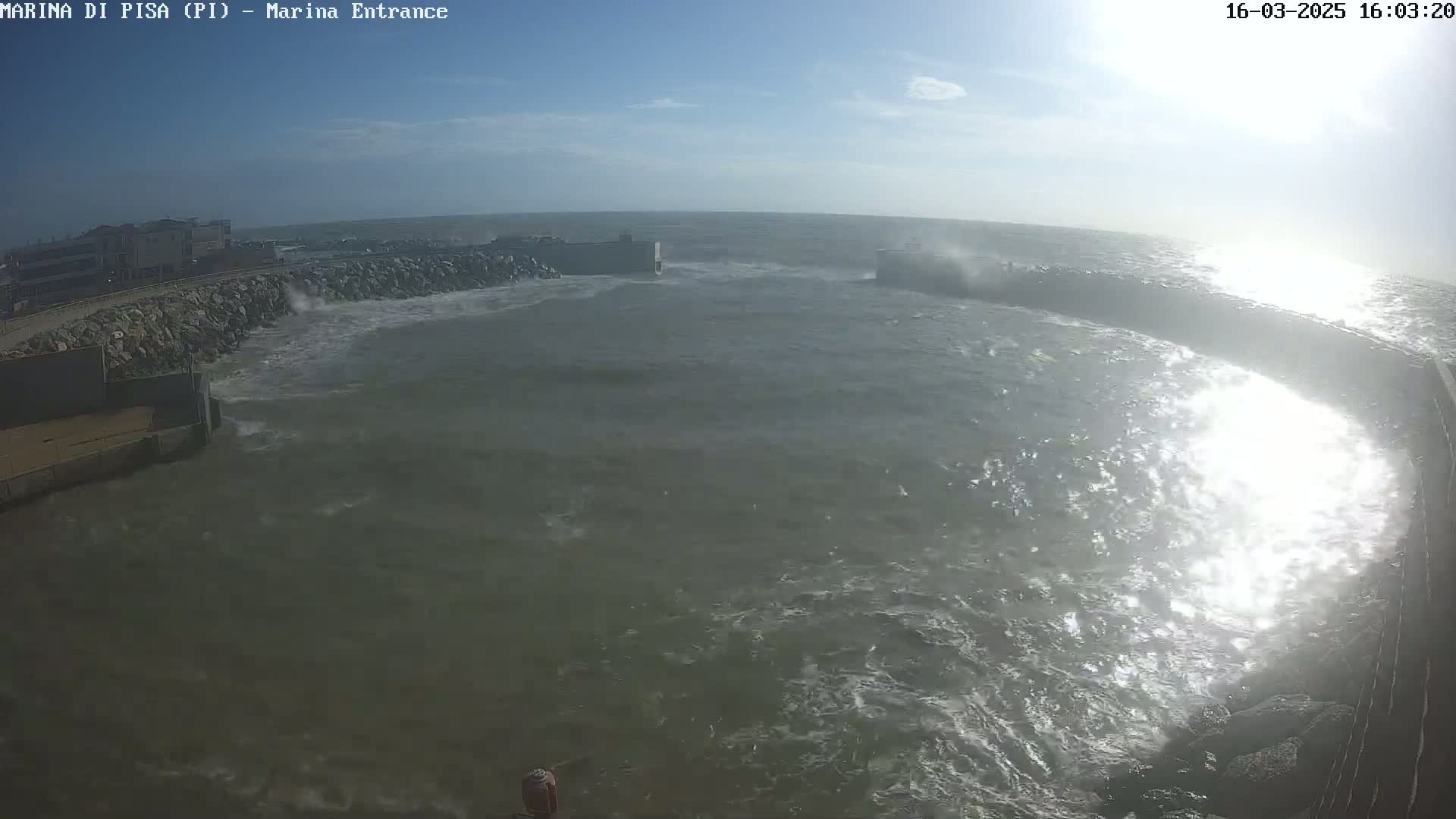 Rough, choppy waves crash against a rocky shoreline and harbor walls under a sunny sky.