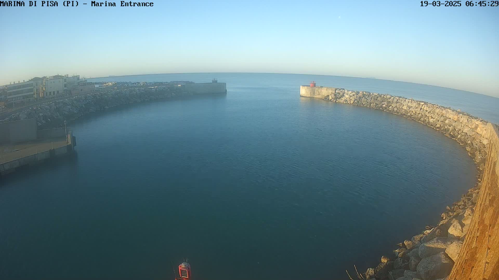 A calm, clear morning reveals a harbor entrance with stone breakwaters curving around a dark-blue body of water, a small red structure visible at one end.