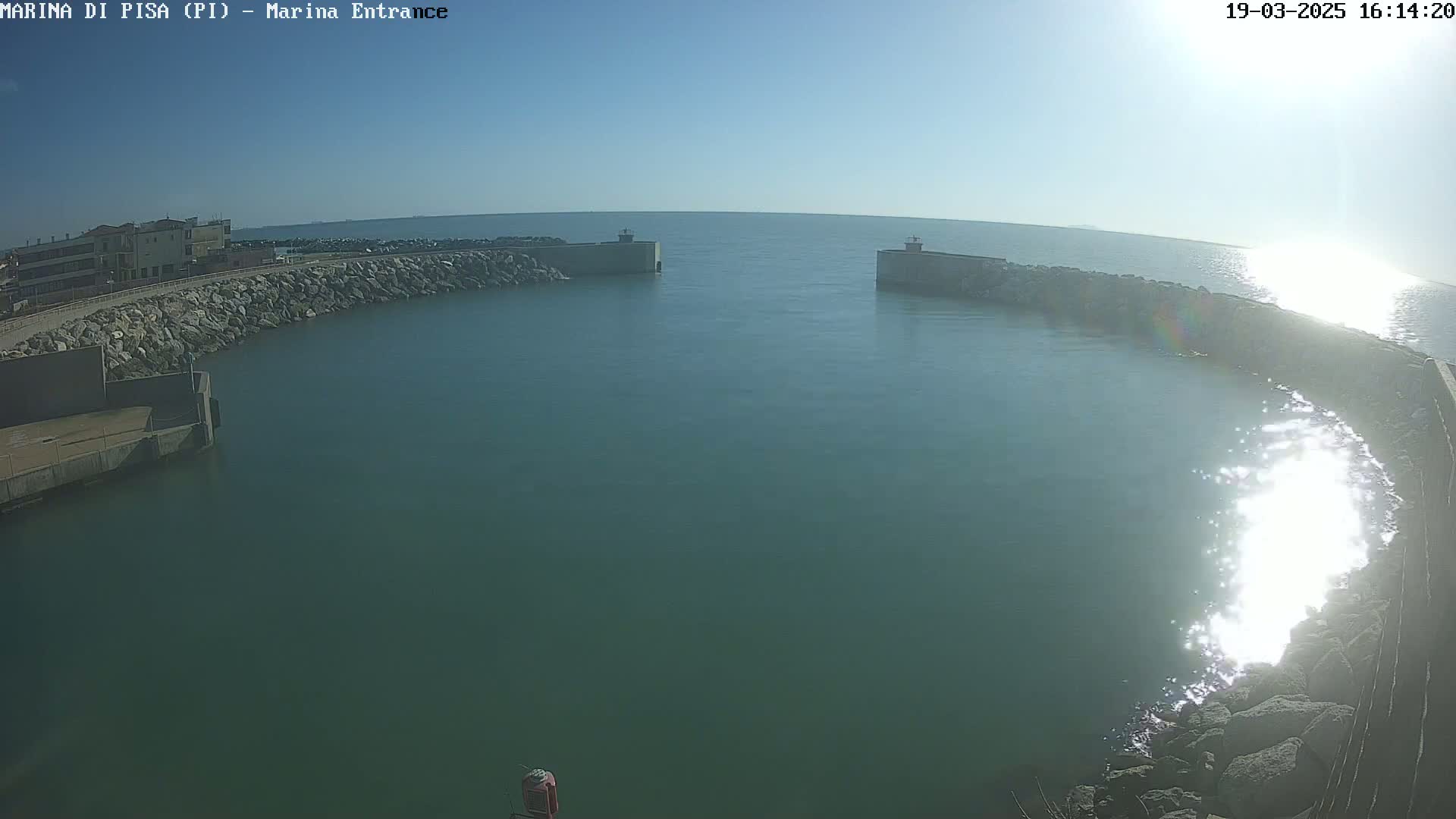 A harbor entrance with calm, sunlit water is seen from an elevated perspective, with rocky breakwaters and a building visible in the background under a clear sky.