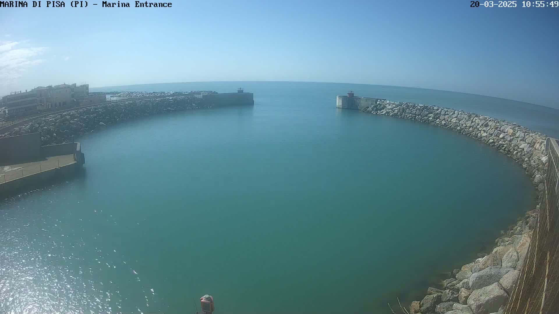 A calm, teal-colored harbor is enclosed by rock walls and buildings under a clear blue sky.