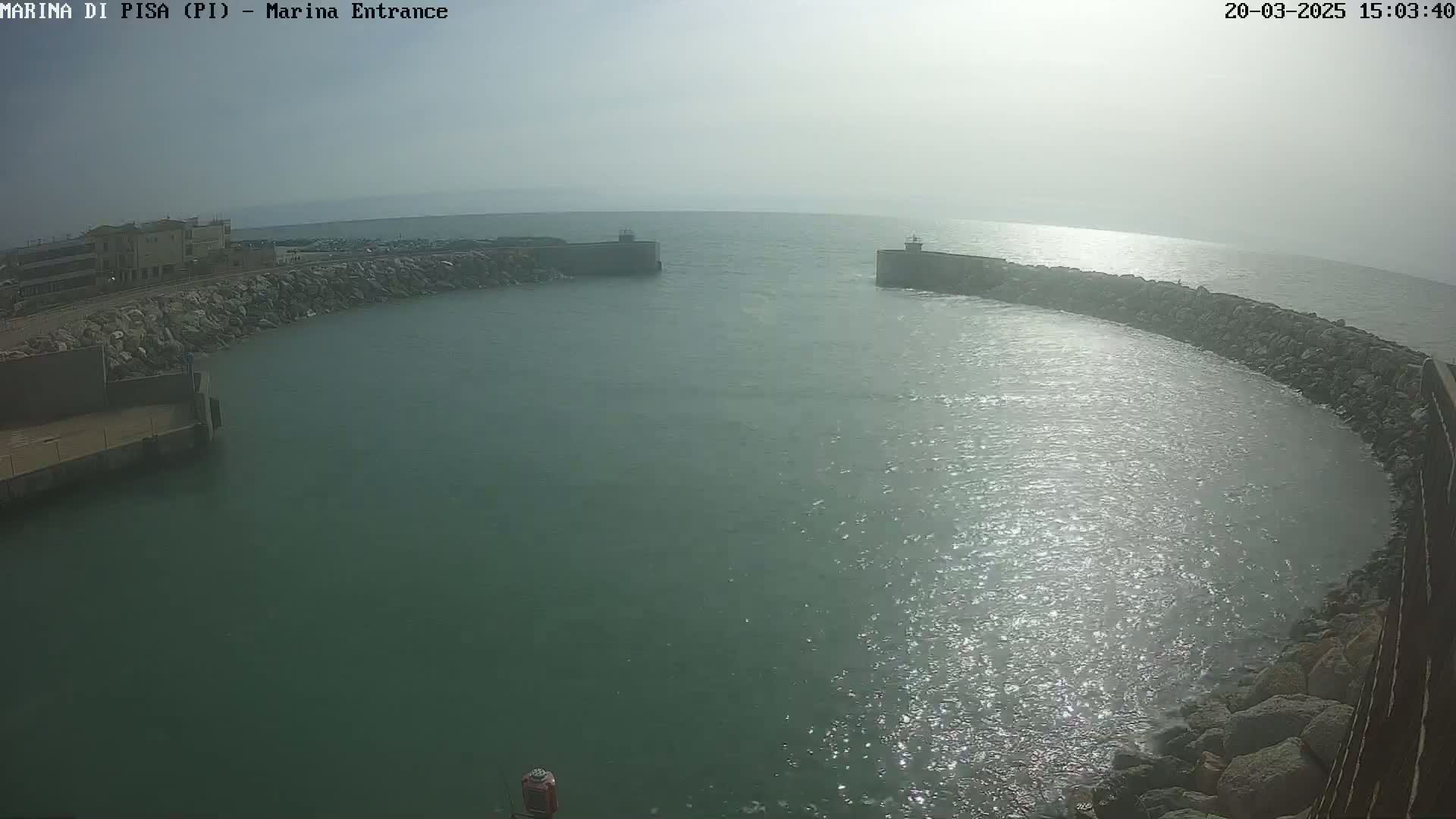 A hazy, sunny day reveals a marina entrance framed by rock walls, with calm, slightly shimmering water filling the harbor.