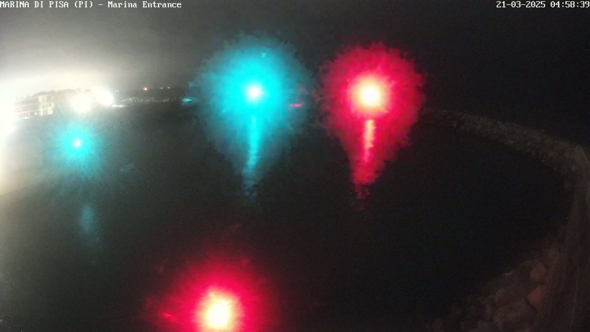 At night, three bright lights, one teal and two red, shine on dark water near a shoreline.