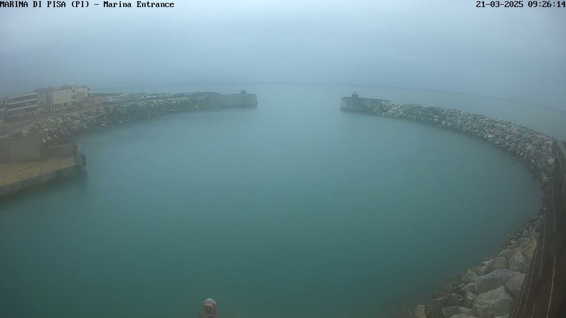 A hazy, foggy view of a harbor entrance with calm, teal water, partially enclosed by rock barriers and buildings.