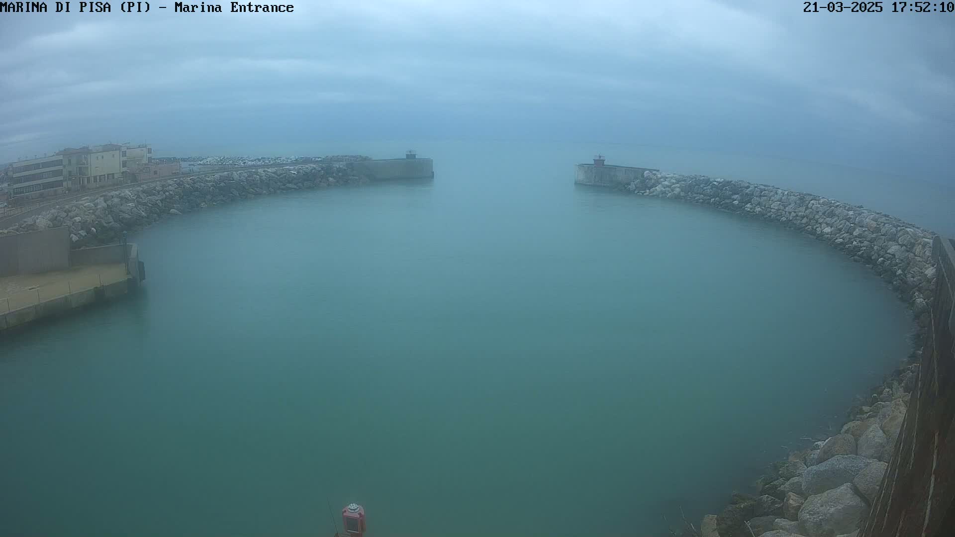 A hazy, overcast day reveals a calm, teal harbor entrance bordered by rock walls and buildings in the distance.