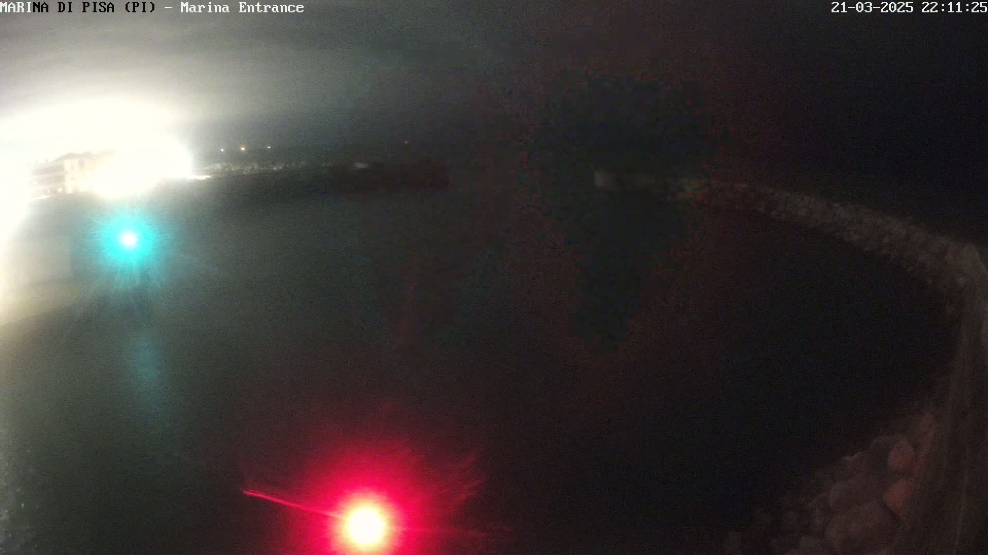 A nighttime, foggy view shows a dark body of water with faint lights on the far shore and two bright lights, one red and one teal, closer to the camera.