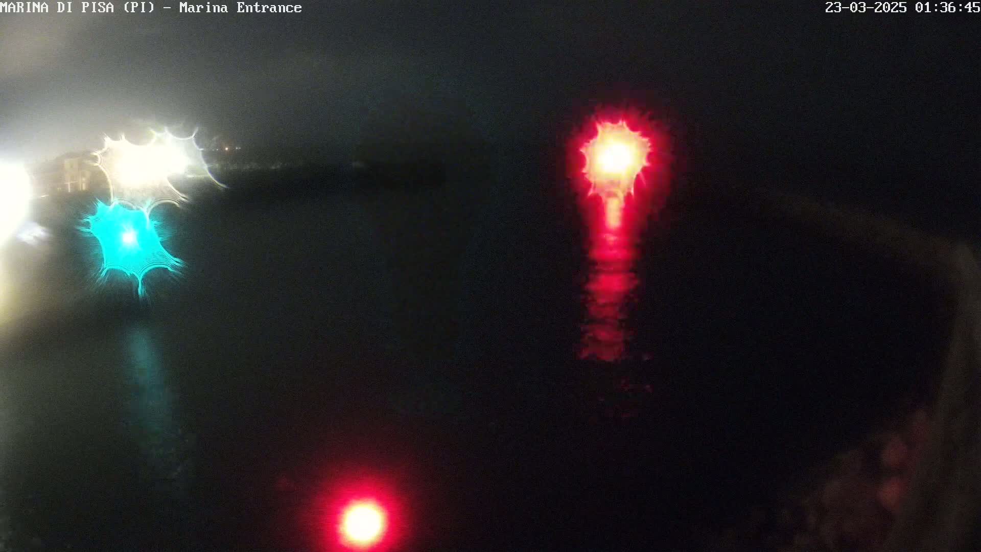 At night, a dark body of water reflects the lights of a nearby structure, including a bright teal light and several red lights.