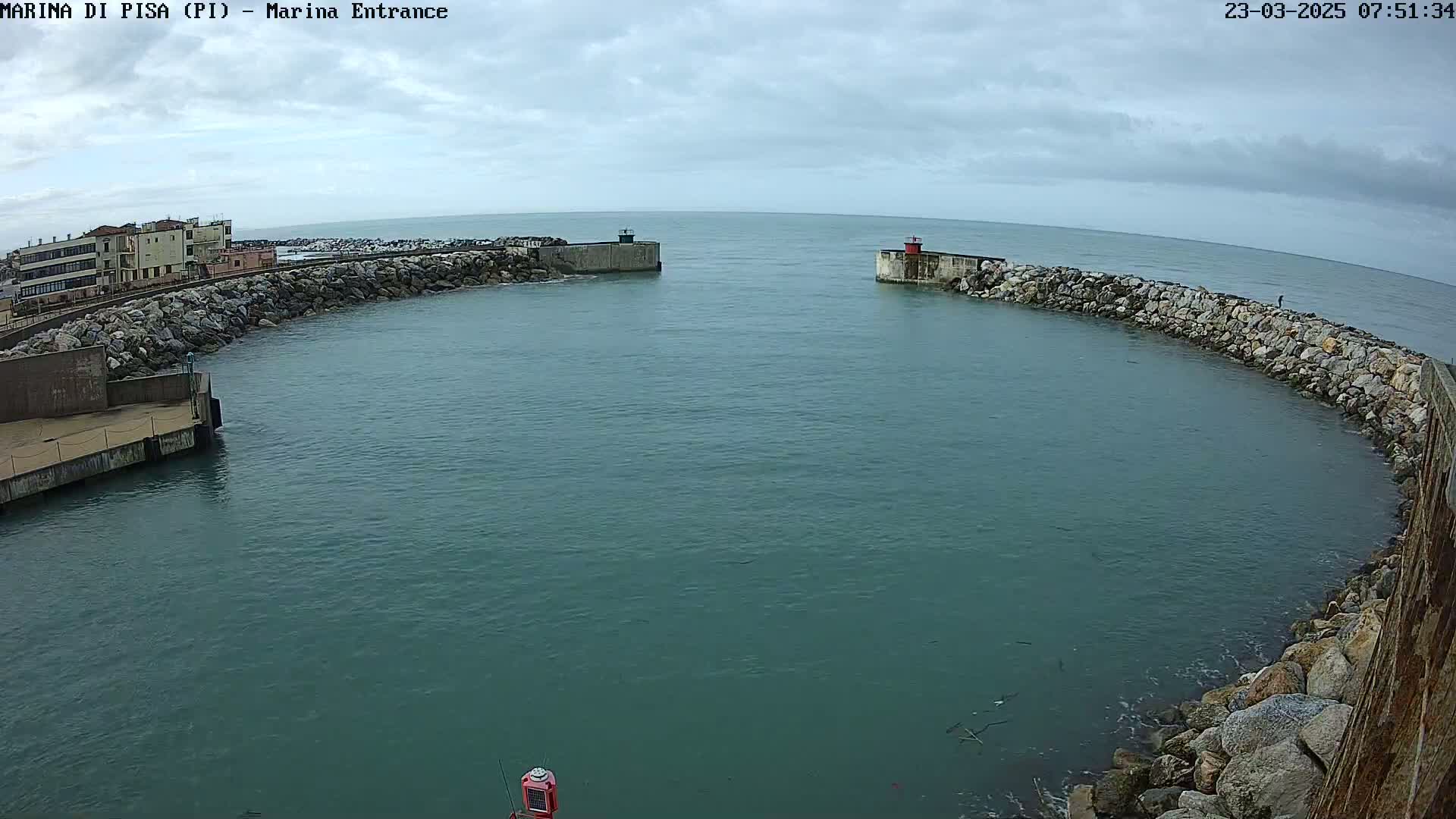 A harbor entrance with calm, teal water is partially enclosed by rock walls and buildings under a cloudy sky.